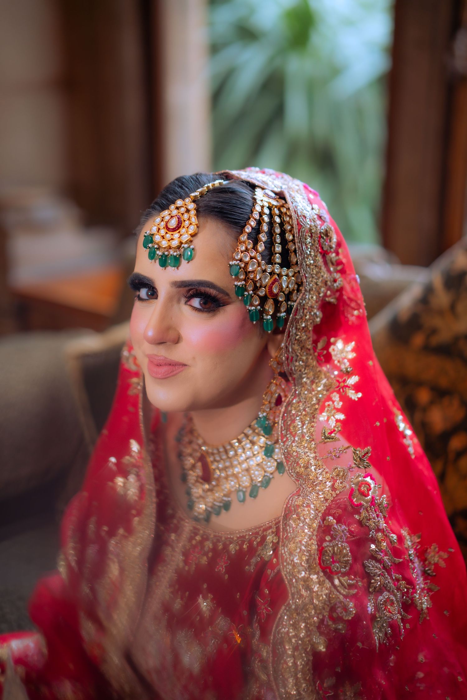 A couple in traditional South Asian wedding attire stand together on a green lawn as the groom adjusts the bride's veil.
