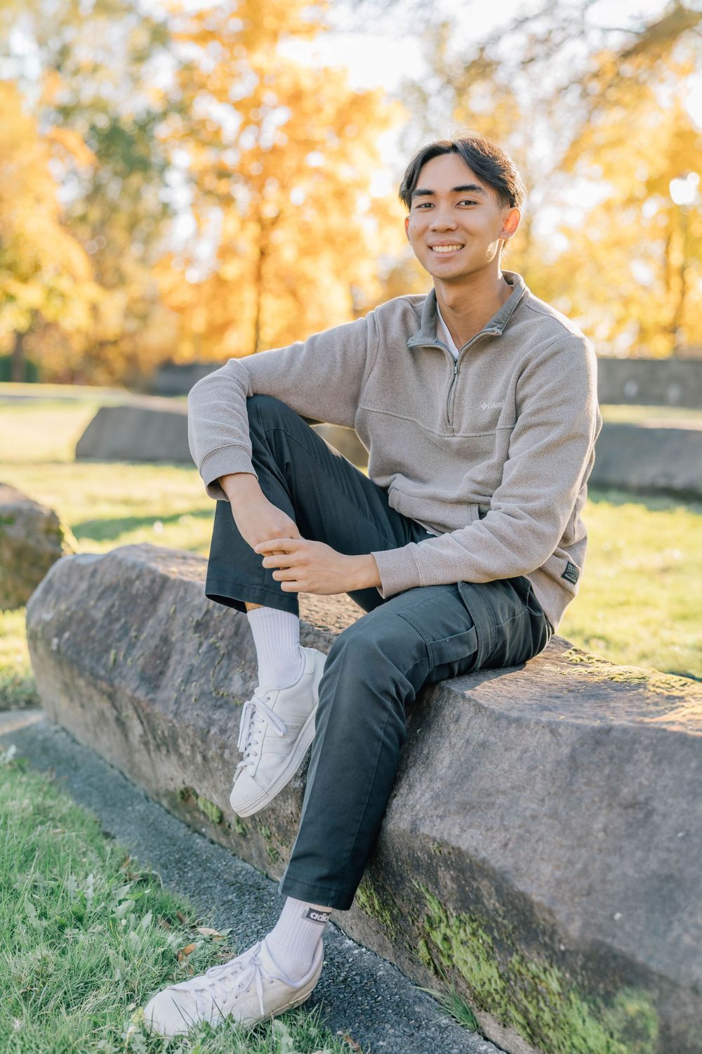 Smiling young Asian man in grey sweatshirt sitting on stone ledge in autumn park setting.