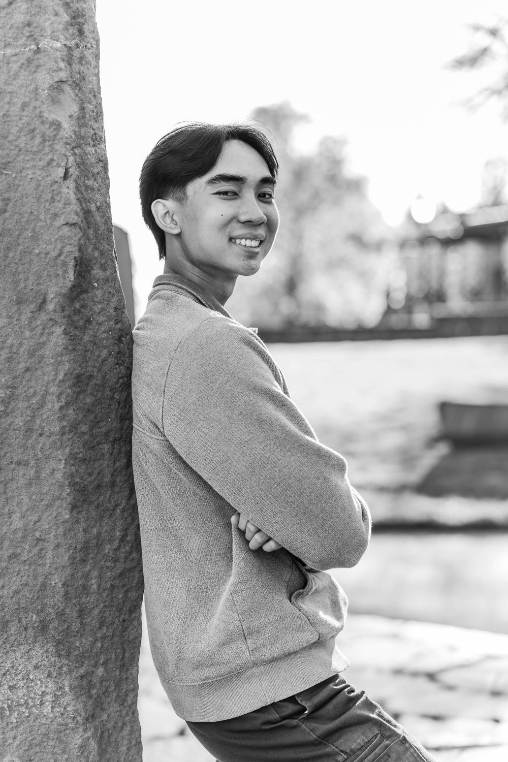 Smiling young Asian man in grey sweatshirt leaning against stone wall in black and white portrait.