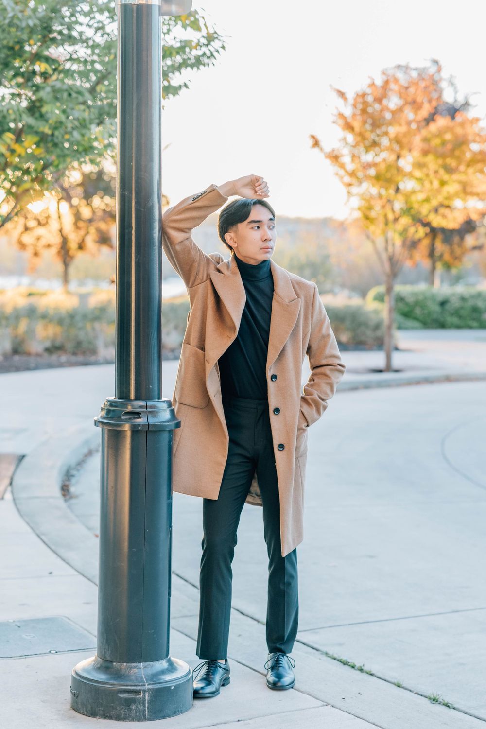 Young Asian man in camel overcoat leaning against lamppost on urban sidewalk with autumn trees.