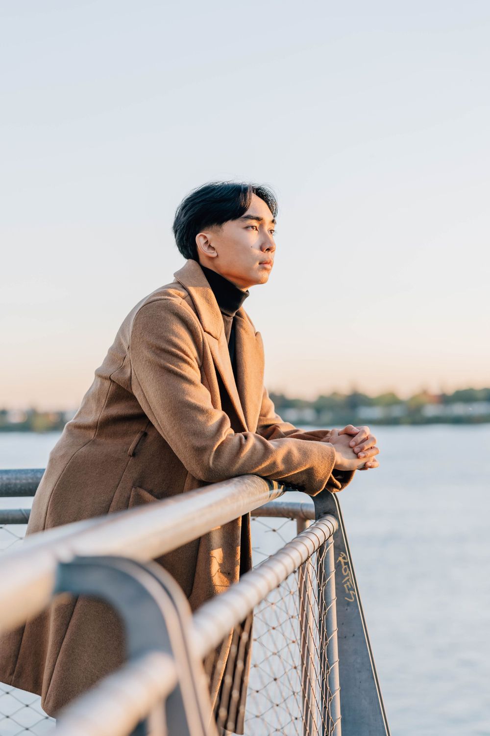 Young Asian man in camel coat leaning on railing gazing at waterfront during golden hour sunset.