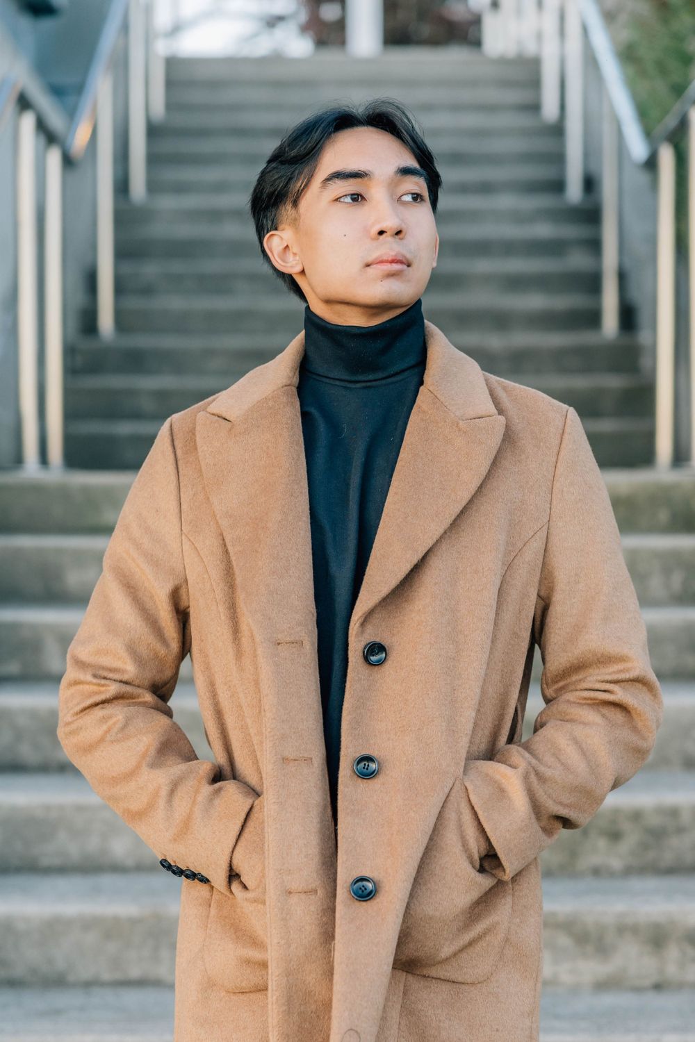 Stylish Asian man in tan overcoat and black turtleneck standing confidently near outdoor staircase.