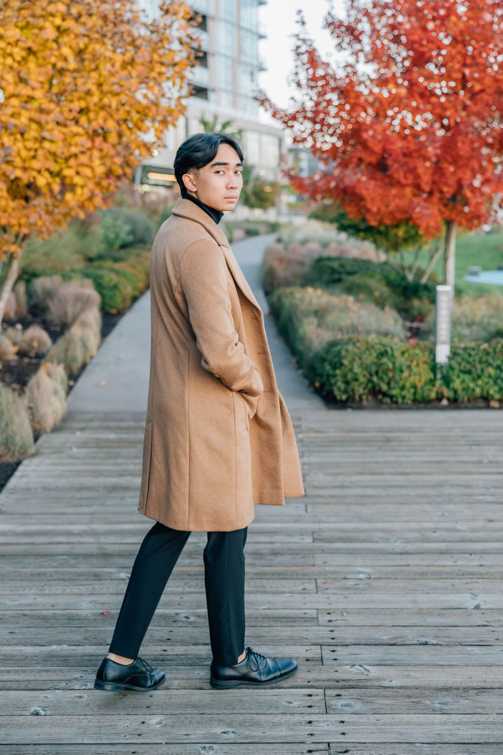 Fashionable Asian man in camel wool coat posing on wooden walkway amid vibrant autumn foliage.