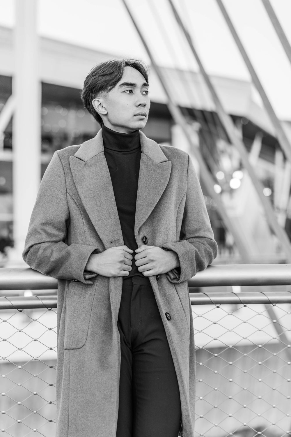 Black and white portrait of young Asian man in long coat adjusting lapels near urban railing.