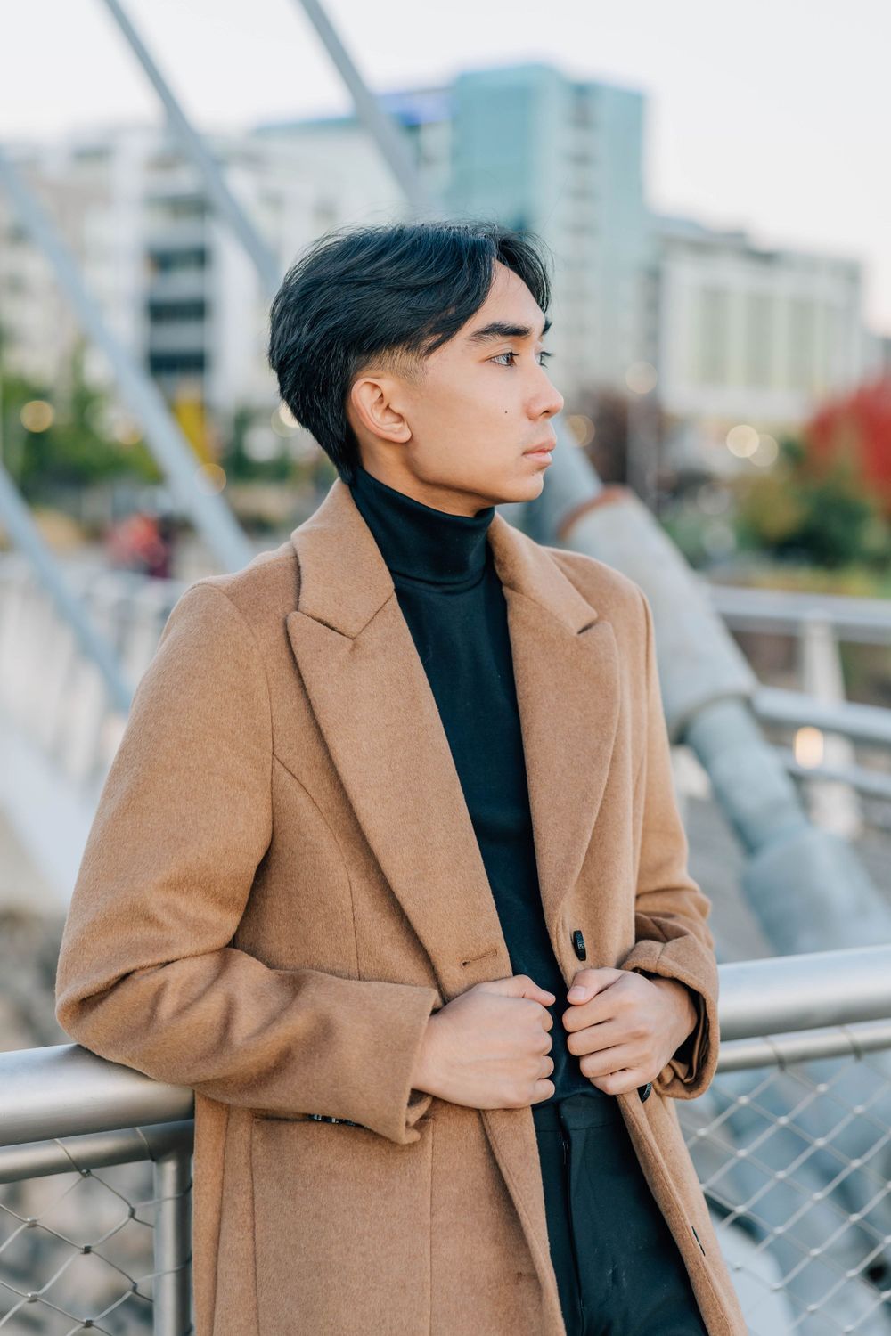 Young Asian man in camel overcoat holding lapels while standing on city bridge at dusk.