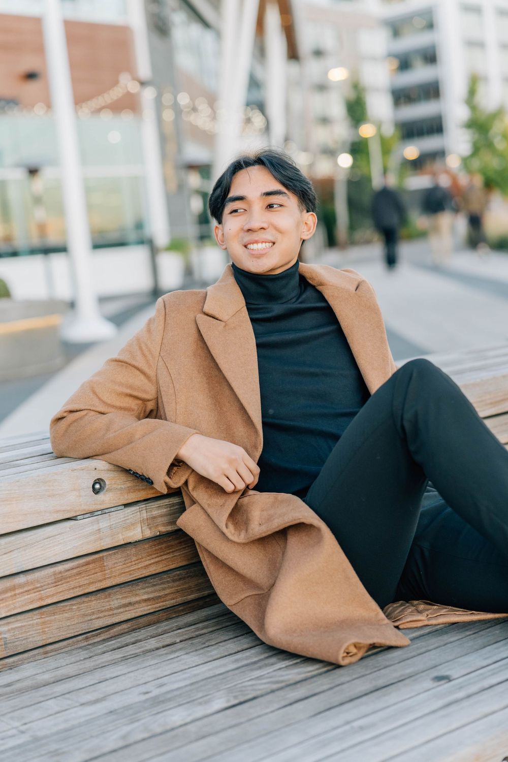 Smiling young Asian man in camel coat relaxing on wooden bench in urban outdoor setting.