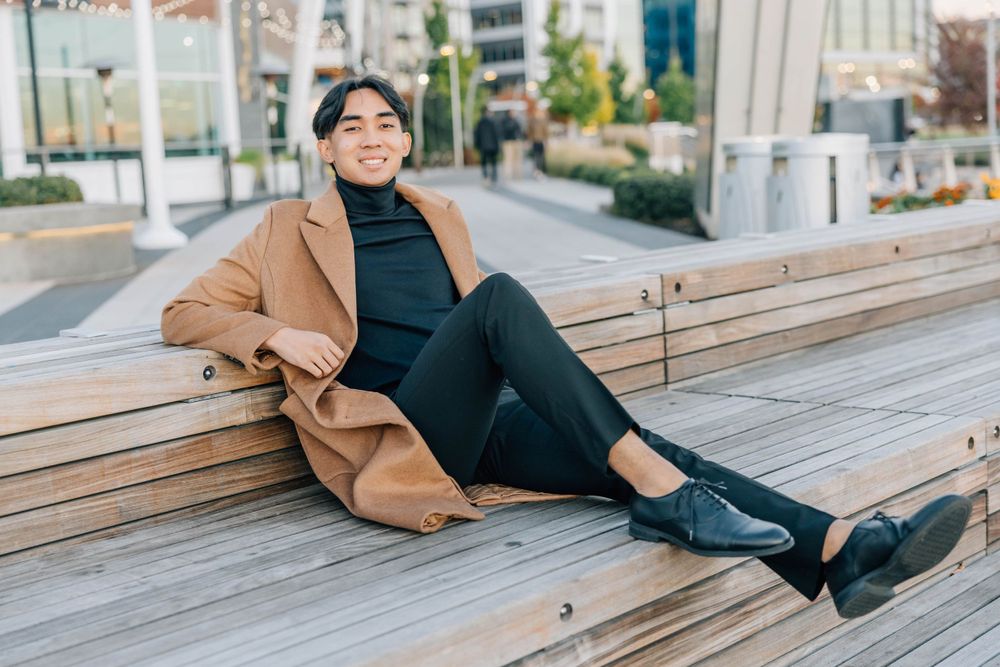 Happy young Asian man in tan coat and black outfit sitting casually on outdoor wooden bench.