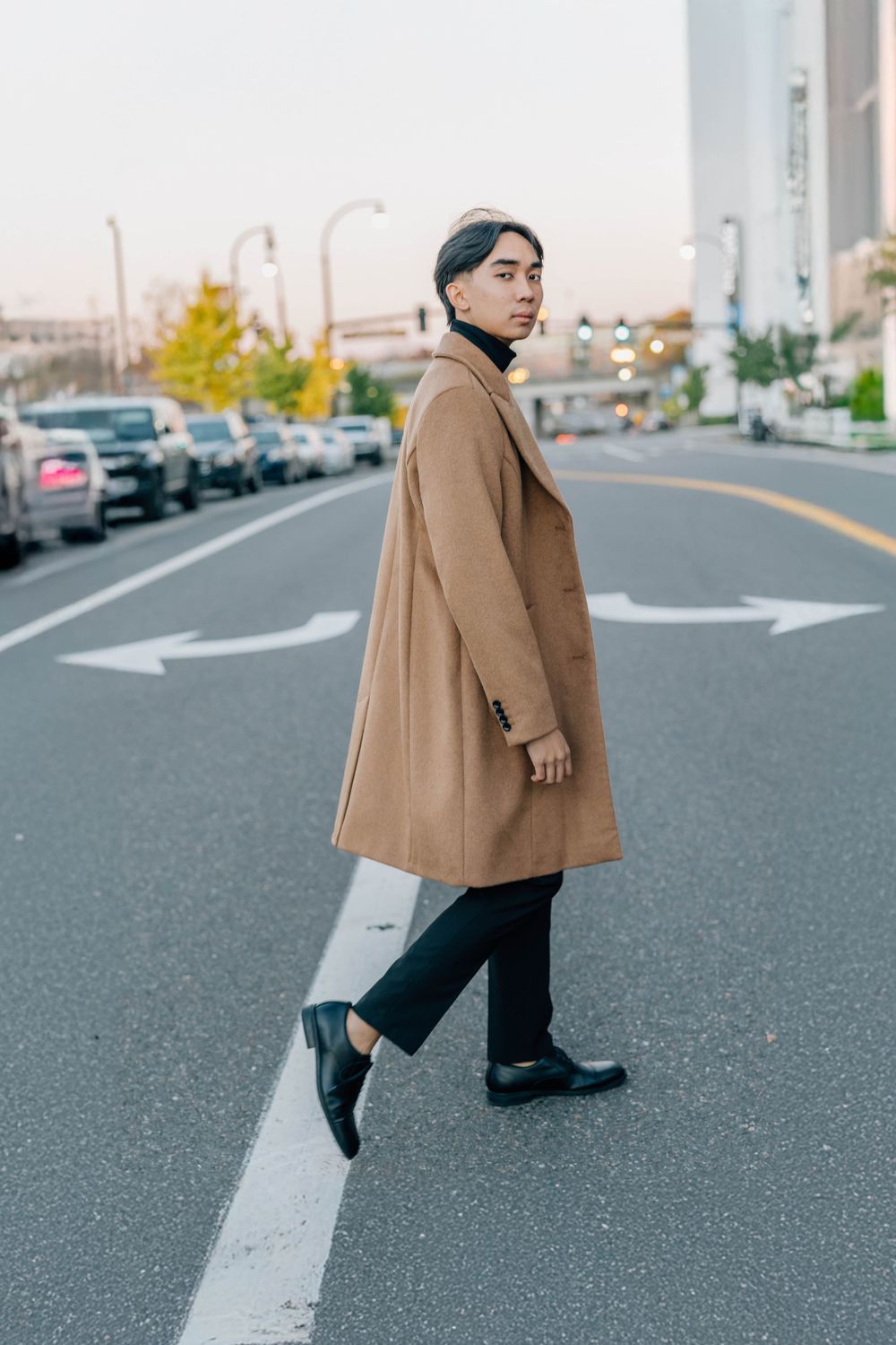 Stylish Asian man in camel overcoat crossing urban street road with arrow markings at dusk.