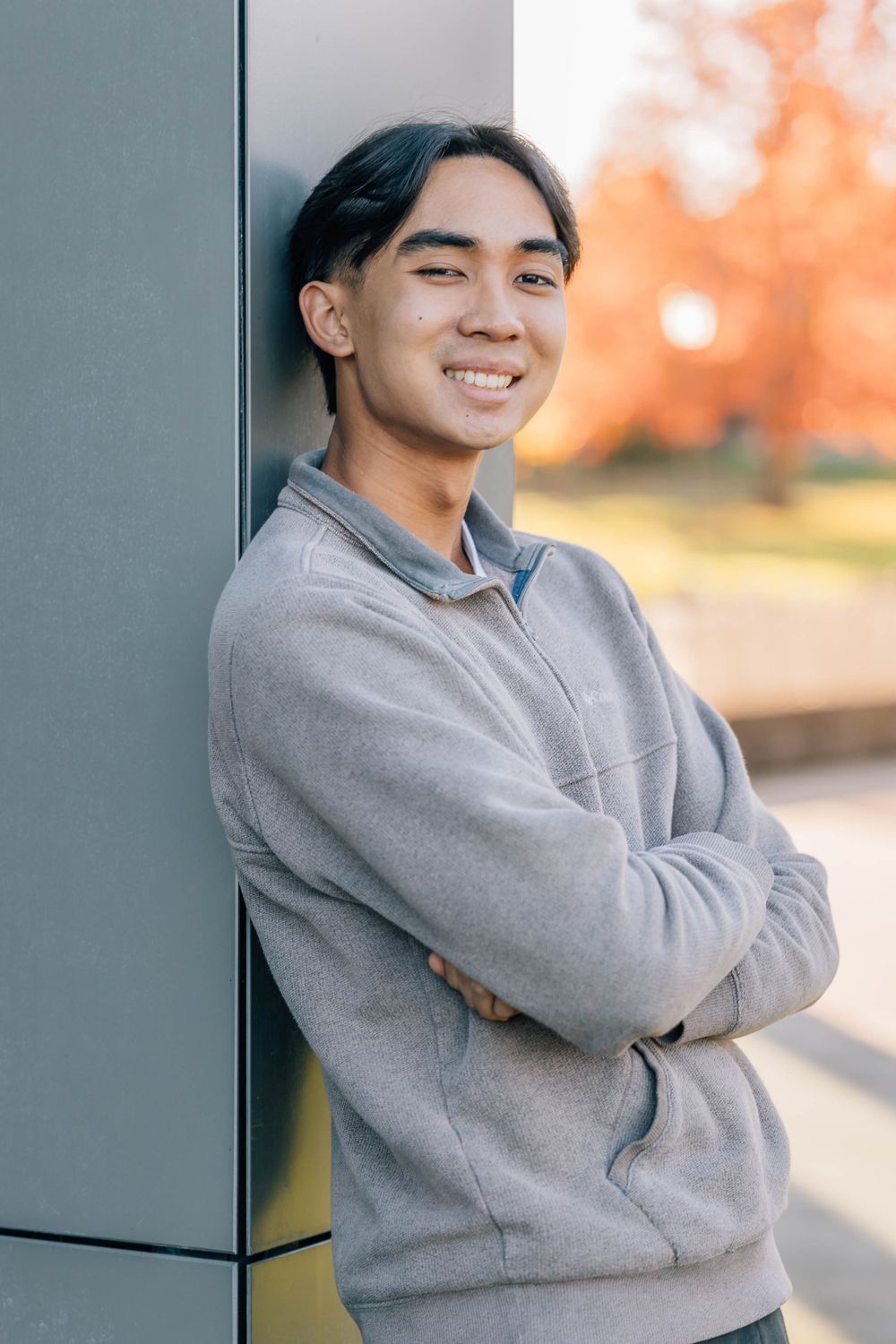 Smiling young Asian man in grey sweatshirt leaning against wall with arms crossed outdoors.