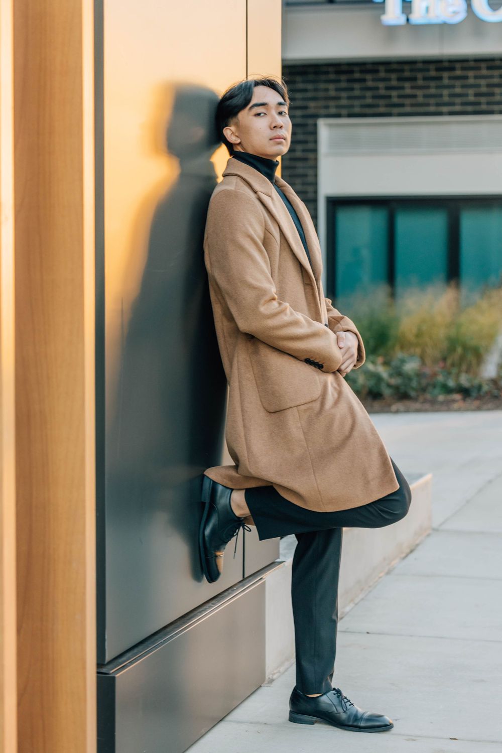 Fashionable Asian man in camel coat leaning against warm-lit wall in urban shopping district.