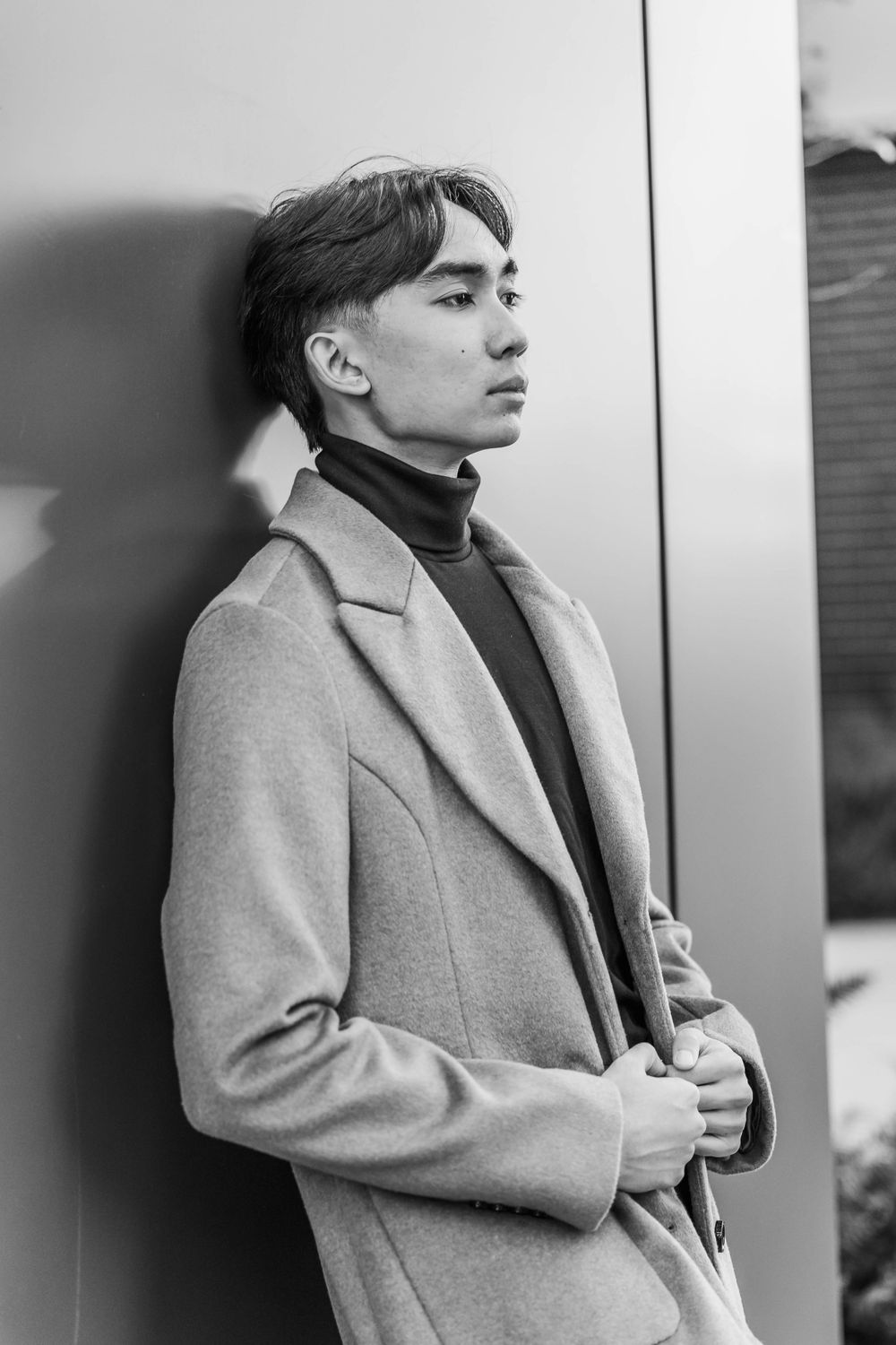 Young Asian man in camel coat and black turtleneck leaning against wall in black and white photo.