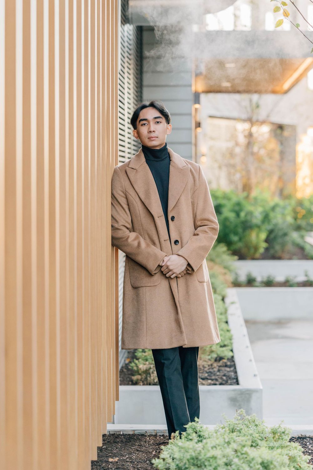 Stylish young man in camel overcoat and black turtleneck posing outdoors against golden wall.