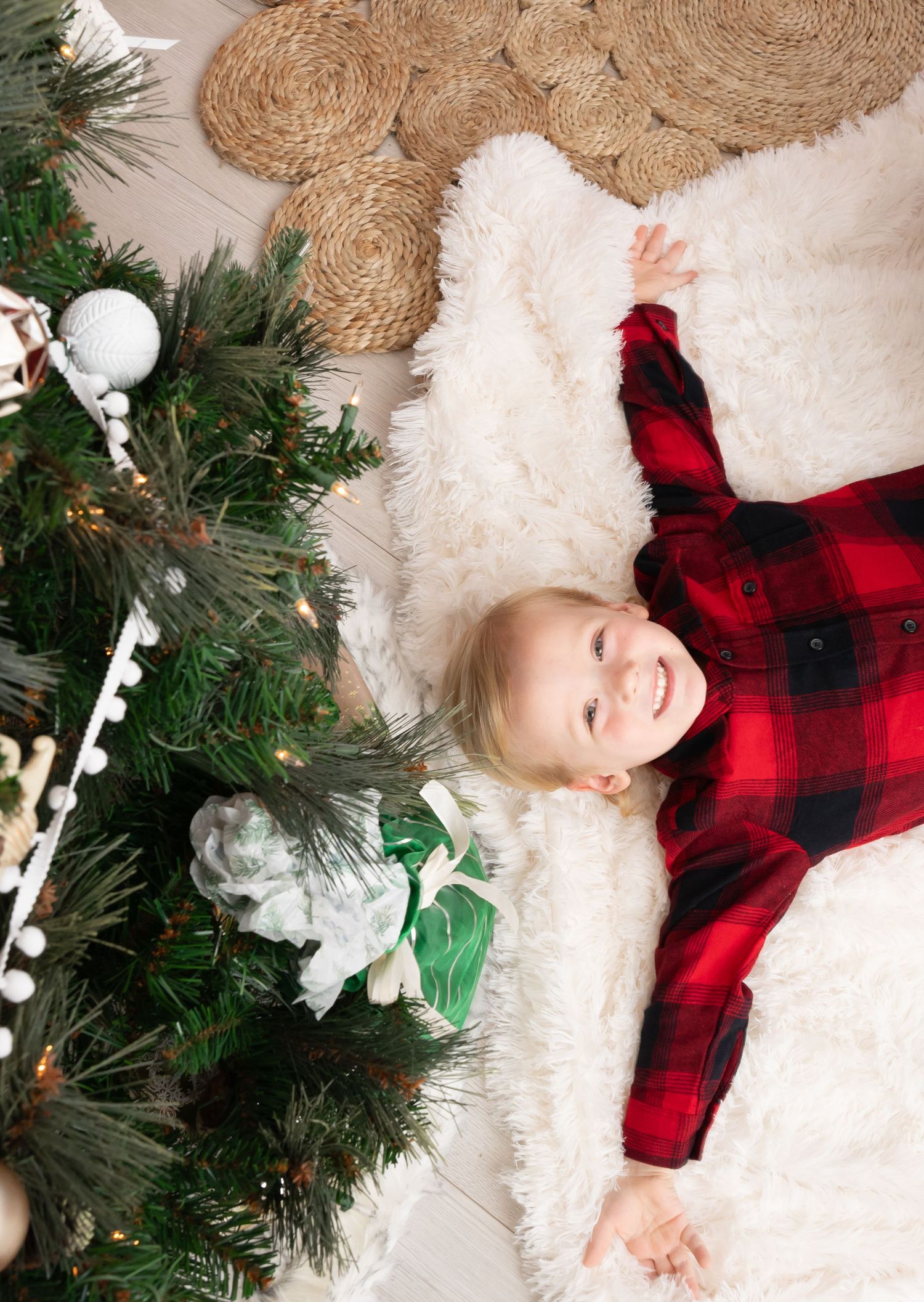 Child laying on white fur rug wearing red plaid next to Christmas decorations.