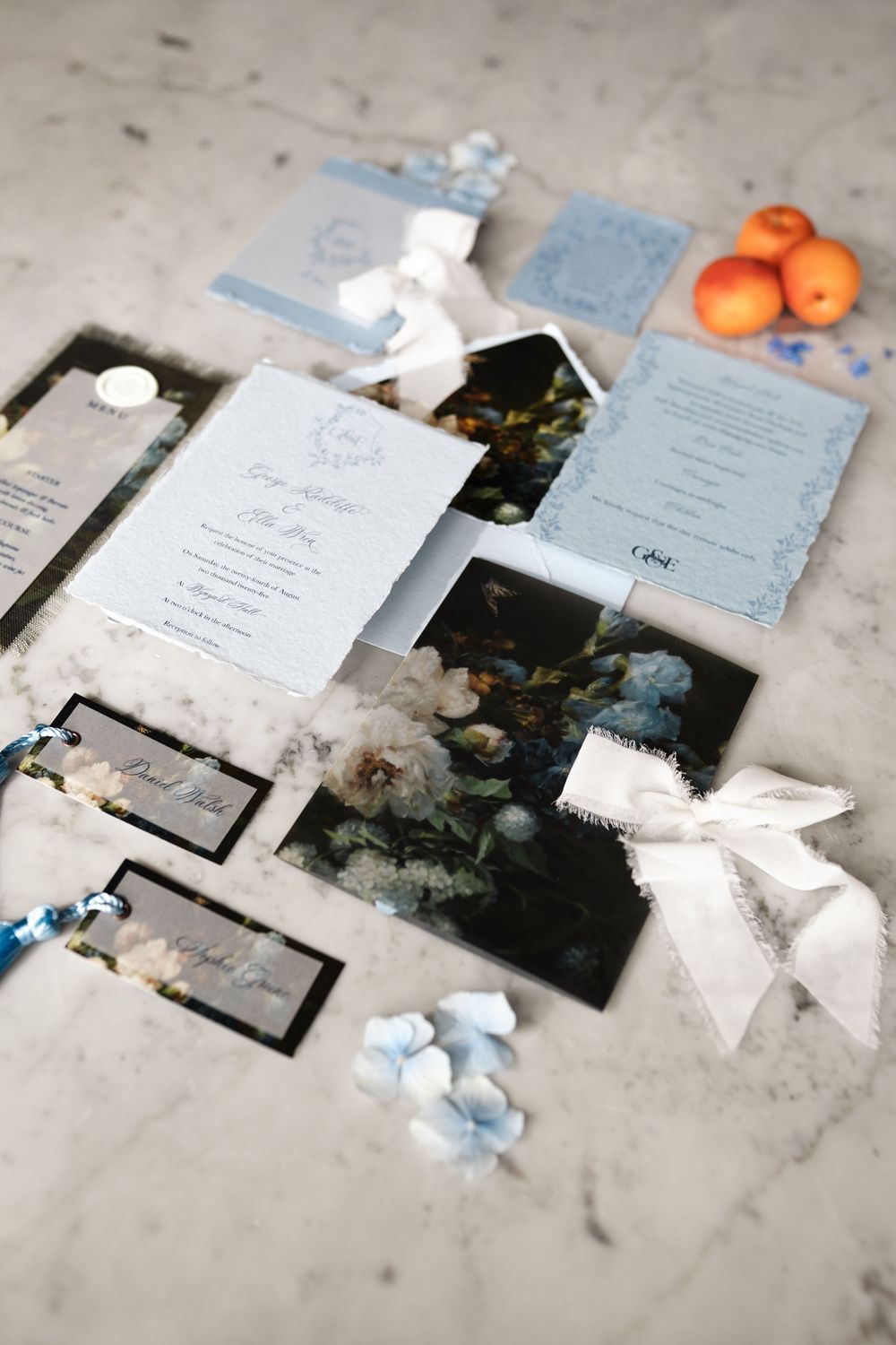 Wedding stationery flat lay with blue and white invitations, ribbons, small flowers, and oranges on a light surface.