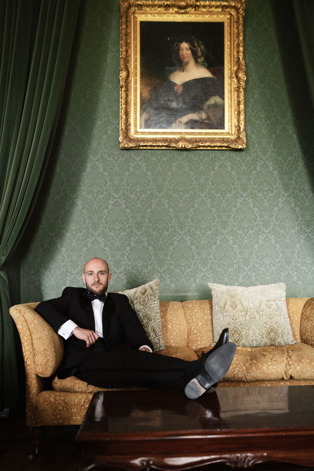 A person in a tuxedo relaxes on an ornate gold sofa beneath a classical portrait in an elegant green-walled room.