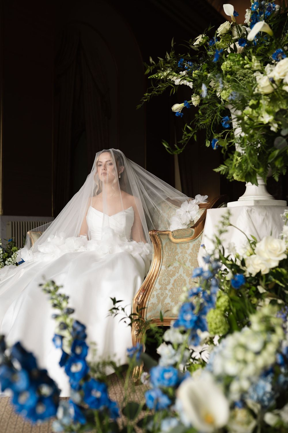 Elegant bridal scene with white and blue floral arrangements in ornate vases framing a classical chair and veil.