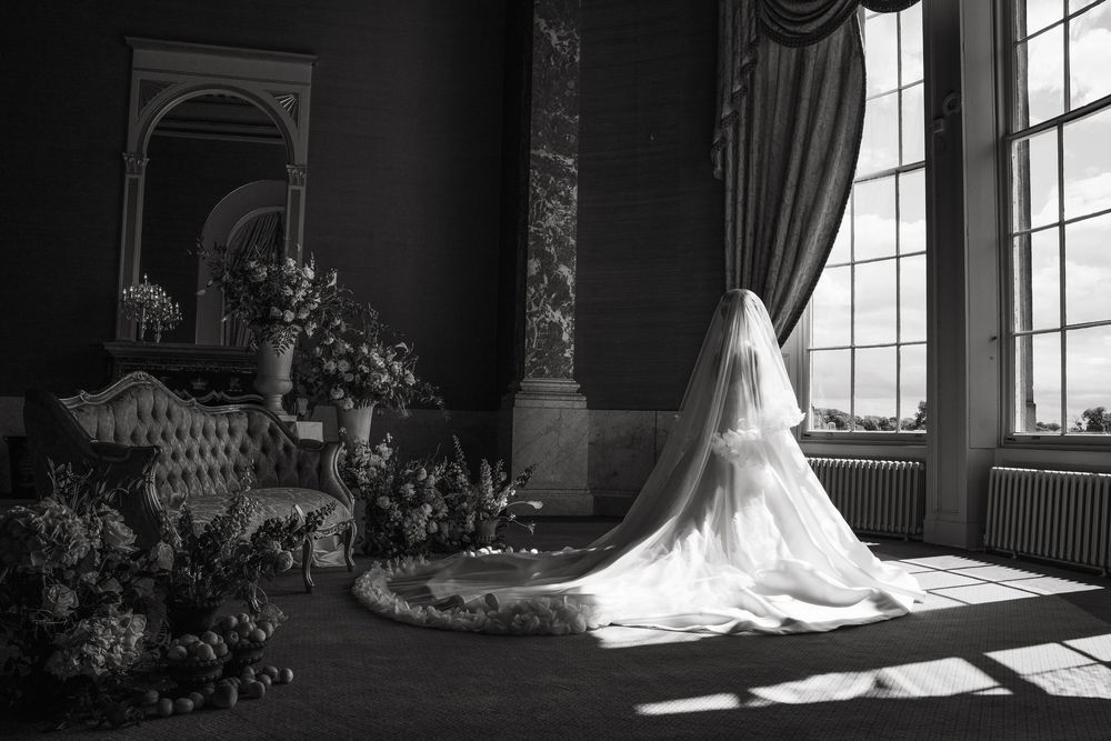 A black and white photograph shows a bride in a flowing white gown standing in an ornate ballroom with sunlit windows.