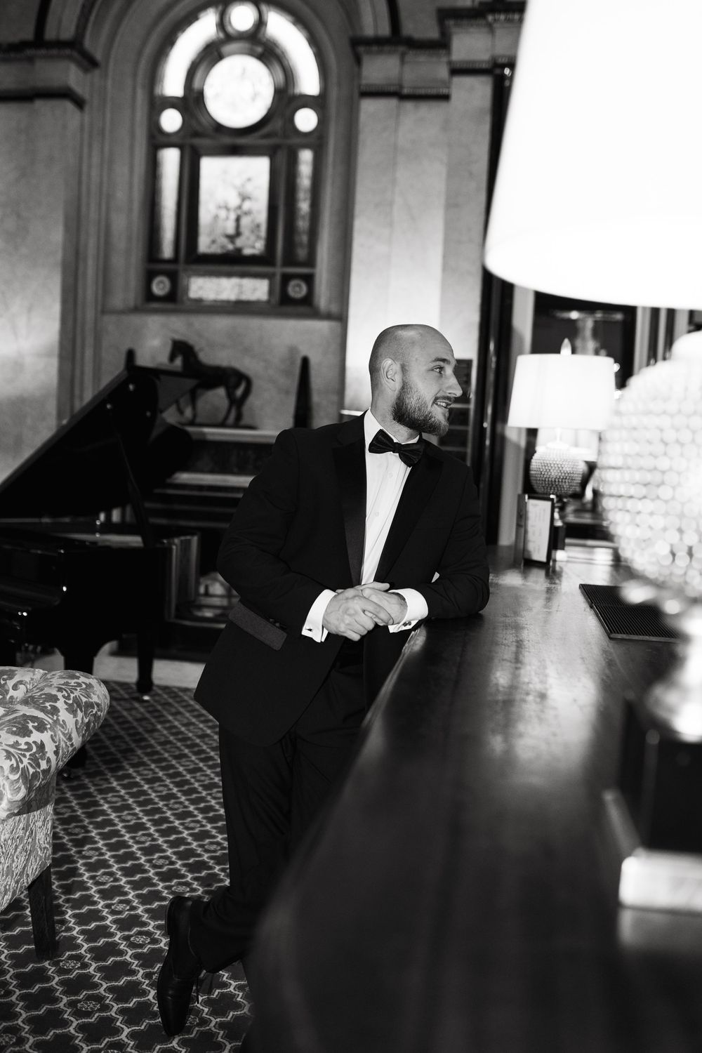 A black-and-white photograph shows someone in a tuxedo seated at a grand piano in an elegant room with an arched window.