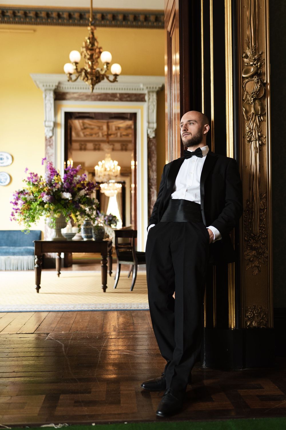 Person in formal black tuxedo and bowtie leans elegantly against ornate doorway in luxury mansion interior.