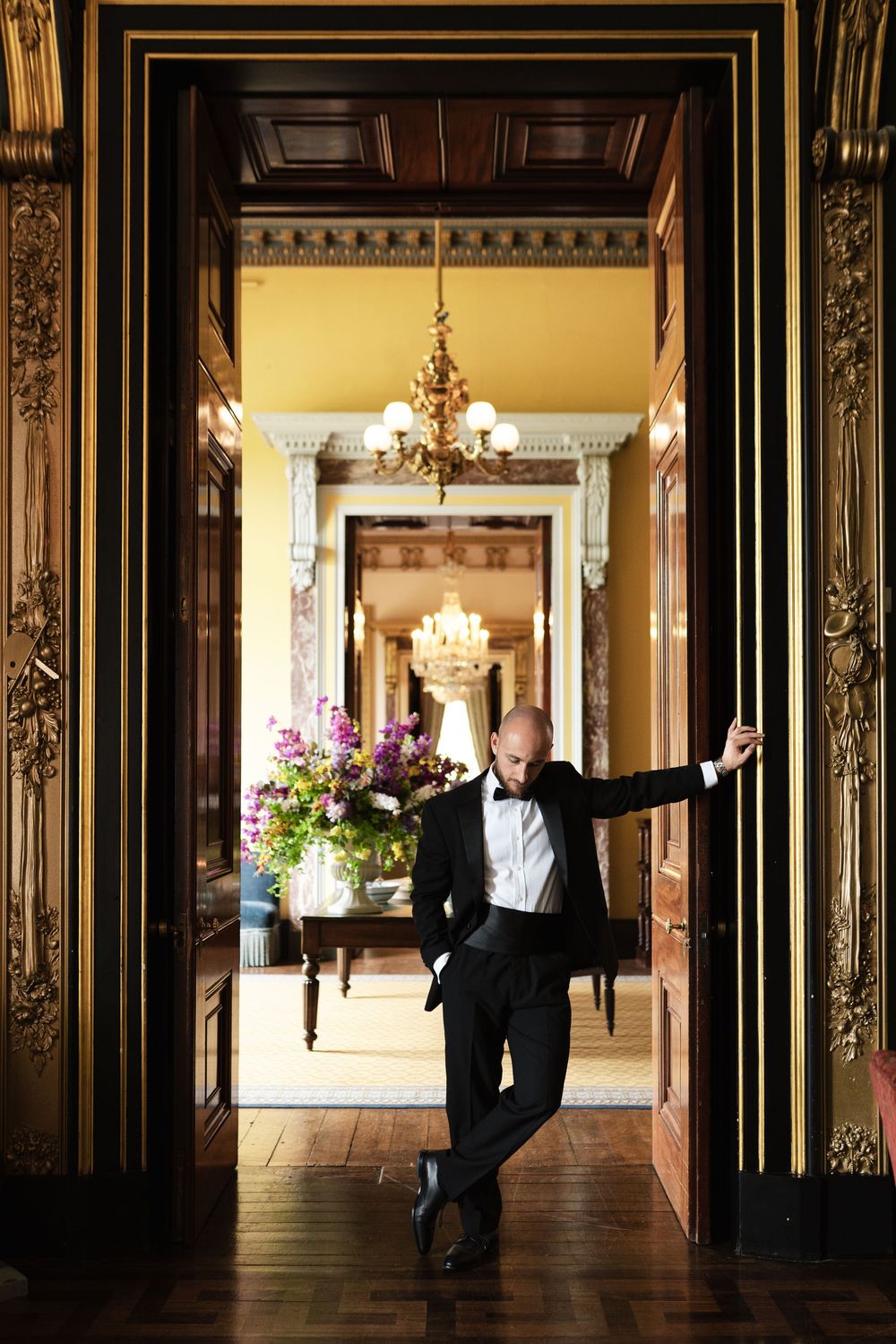 Elegant ballroom entrance with ornate gold doorframe, crystal chandeliers and floral arrangements in luxurious mansion interior.
