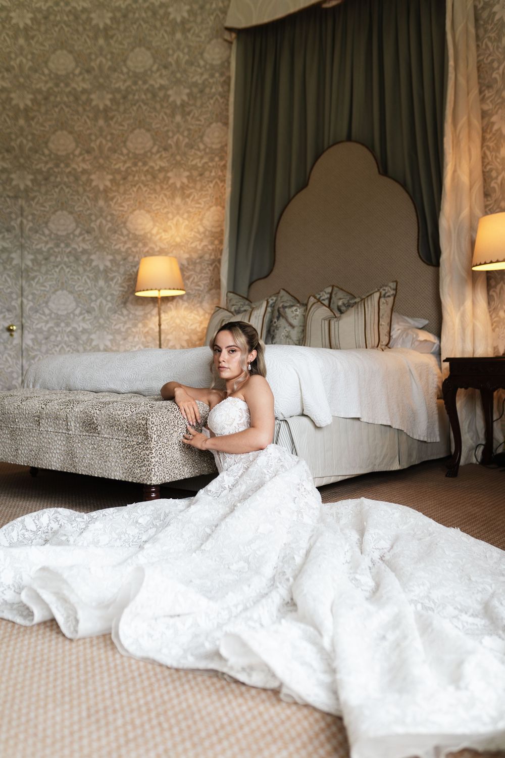 A white lace wedding dress flows across beige carpet in an elegant bedroom with ornate wallpaper and vintage furnishings.