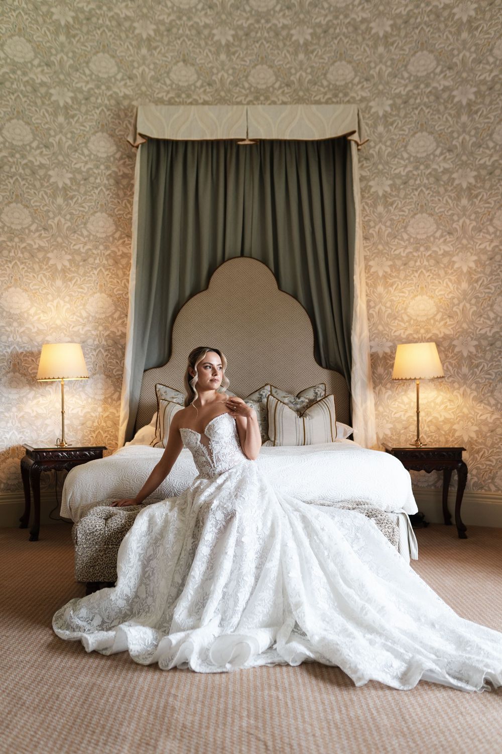 A wedding dress spread dramatically across a luxurious hotel bed with matching table lamps on both sides.