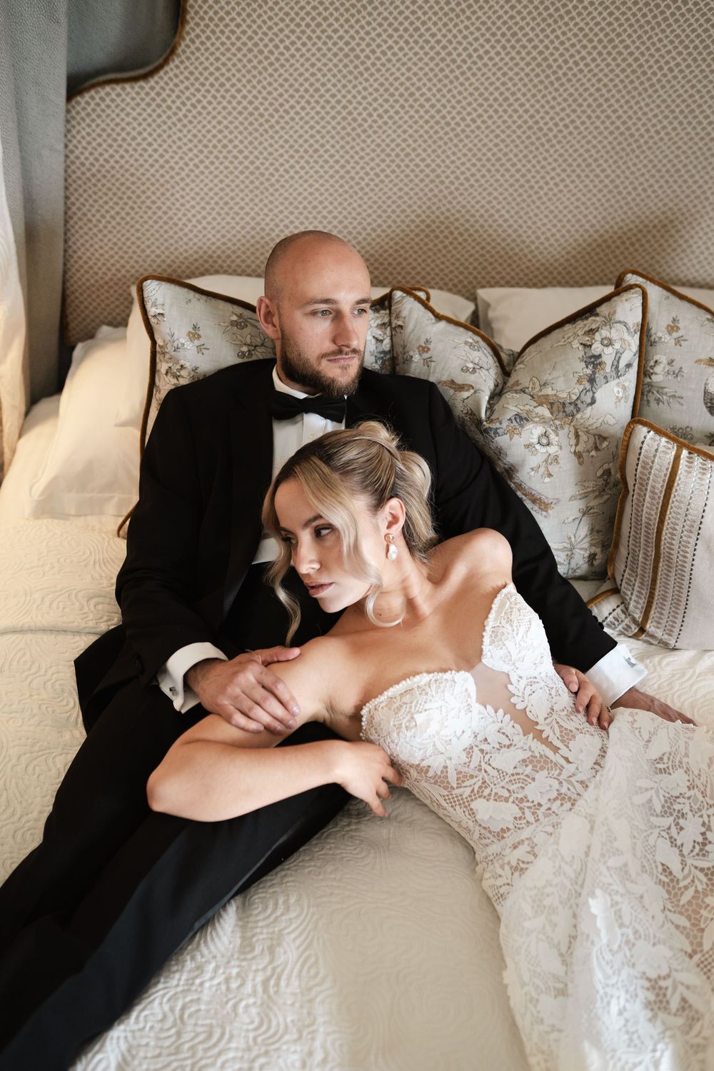 A romantic wedding couple shares tender moments resting together on a beige upholstered loveseat while posing for photos.