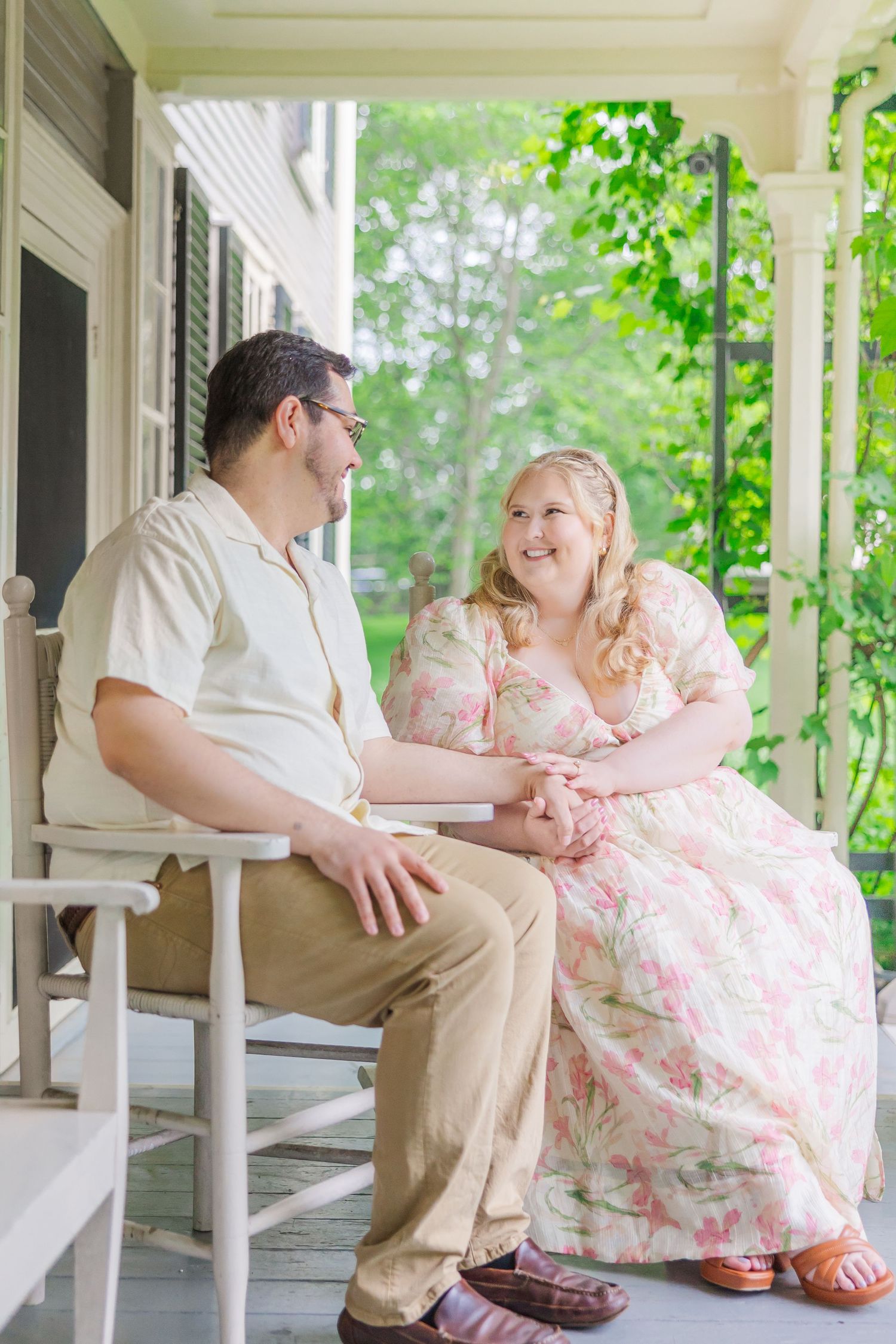 A couple dressed in white and floral attire share a playful moment on a sun-lit front porch with white columns and greenery.