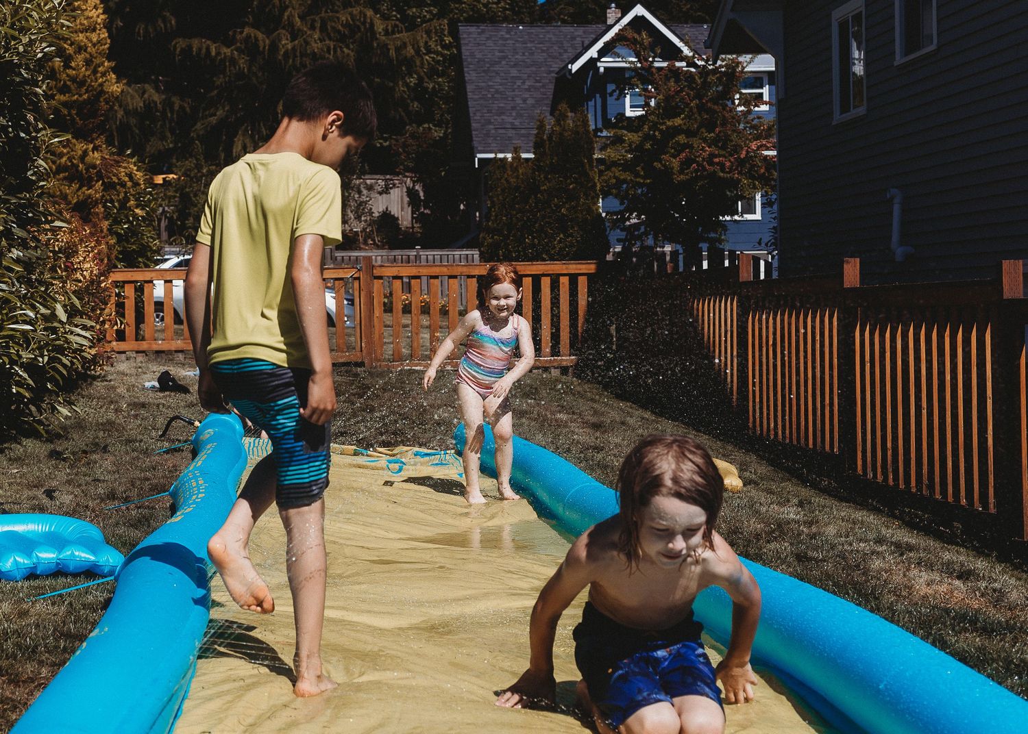 Children sliding down a long blue slip-n-slide on a sunny summer day in a backyard with wooden fencing.