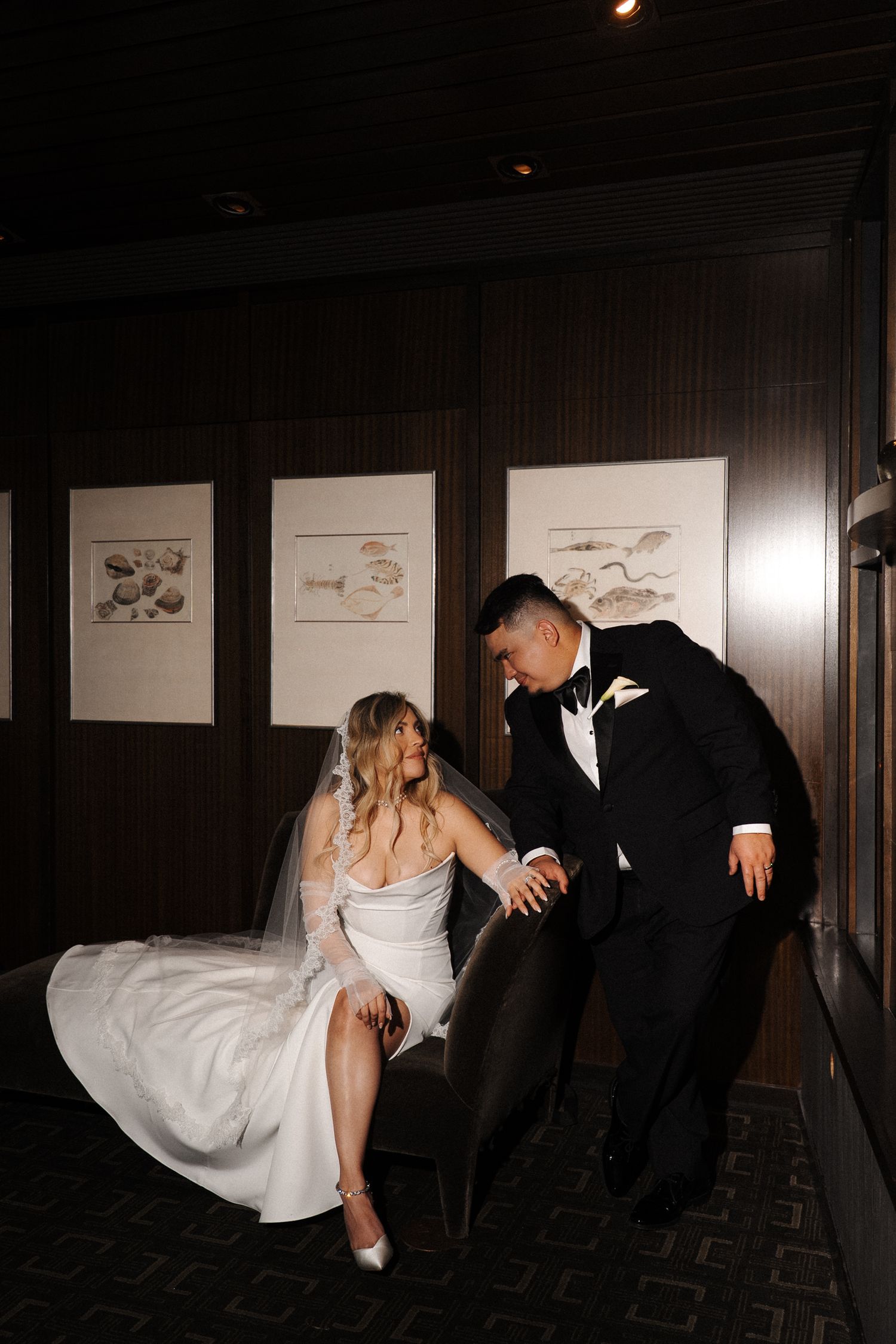 A couple shares a playful moment in formal attire, with the bride lifting her dress to show her shoes against dark walls.