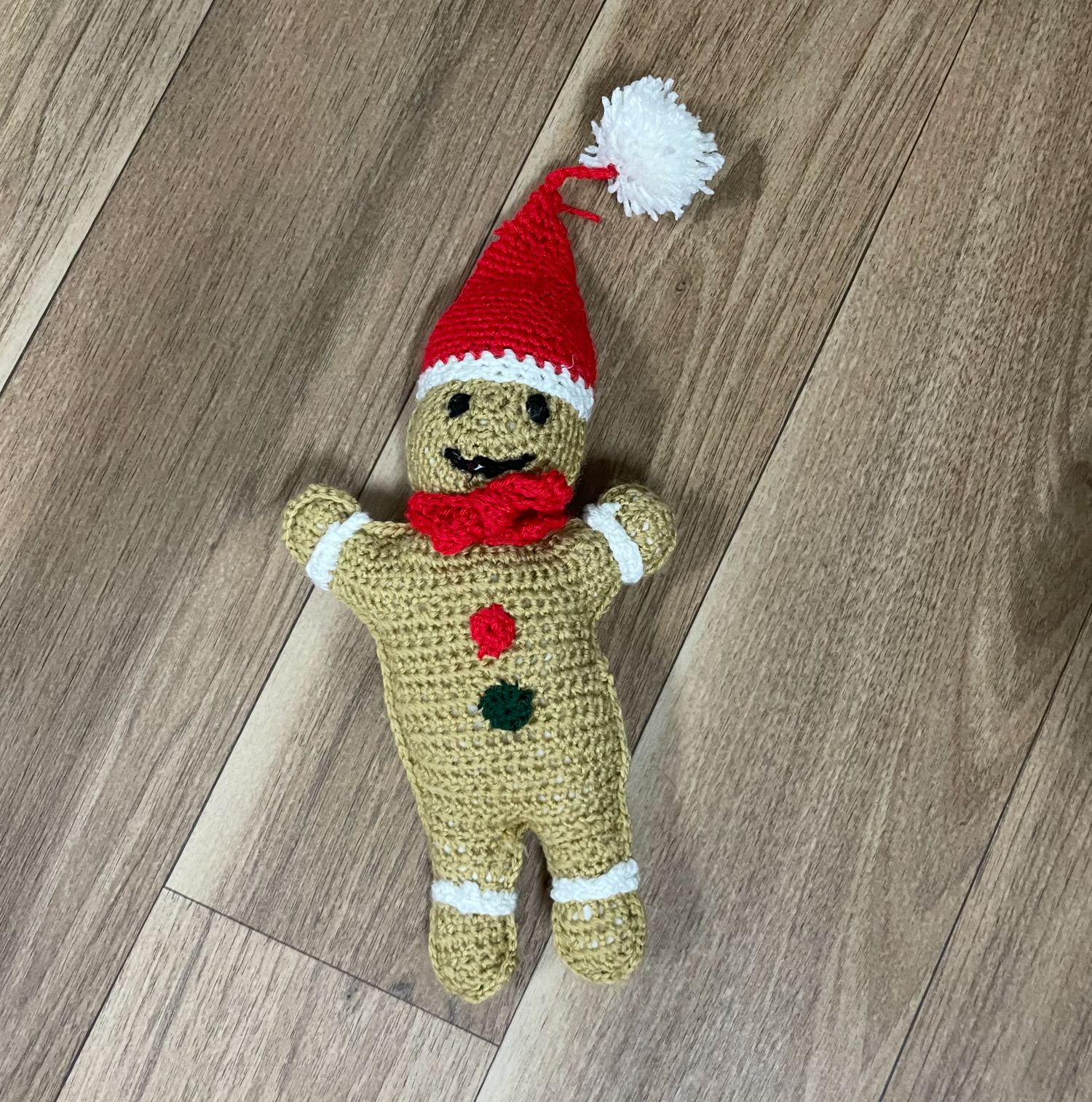 A crocheted gingerbread man doll wearing a red and white Santa hat on wooden flooring.
