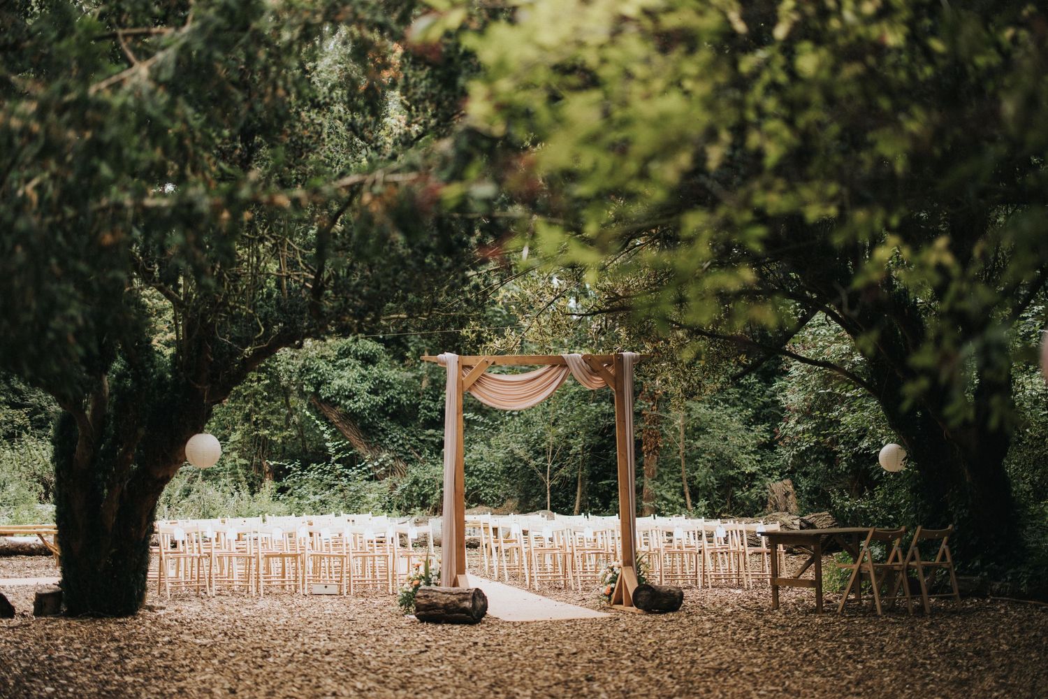 A rustic outdoor wedding ceremony setup with wooden chairs and a floral arch in a wooded garden setting at Hirst Priory.