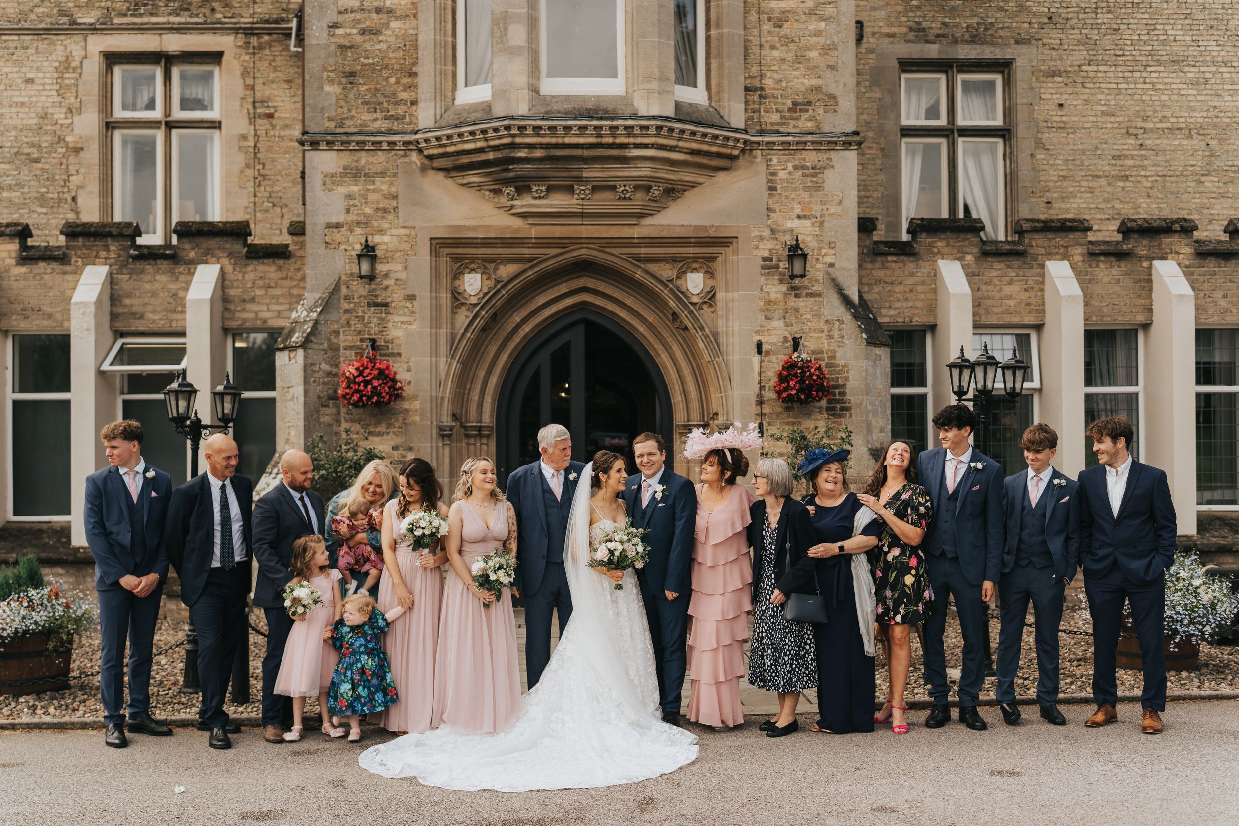 Wedding party poses in front of elegant stone building with arched entrance and hanging flower baskets at Cave Castle.