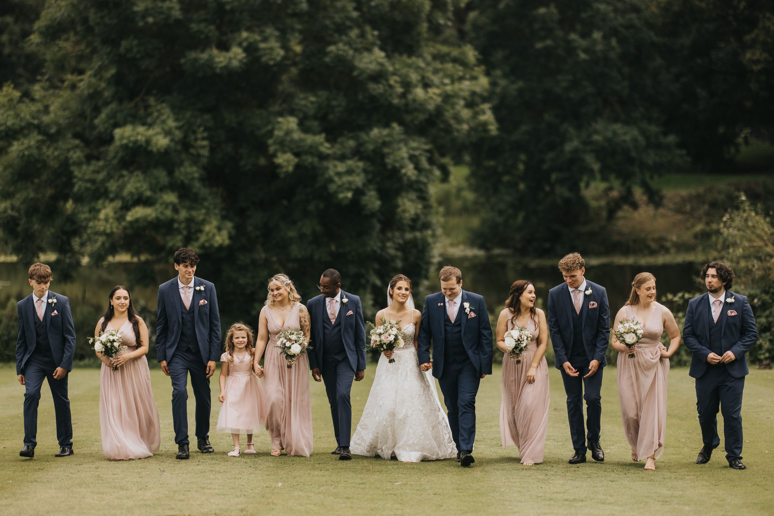 A wedding party in formal attire walks in line on green grass with navy suits and blush pink dresses at Cave Castle.