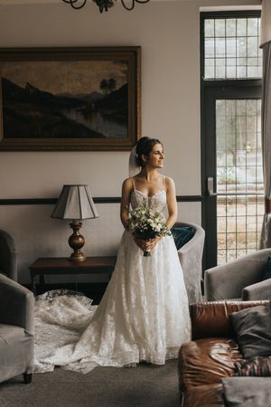 A bride in a flowing white lace dress poses elegantly against a room with vintage decor and brown leather furniture at Cave Castle.