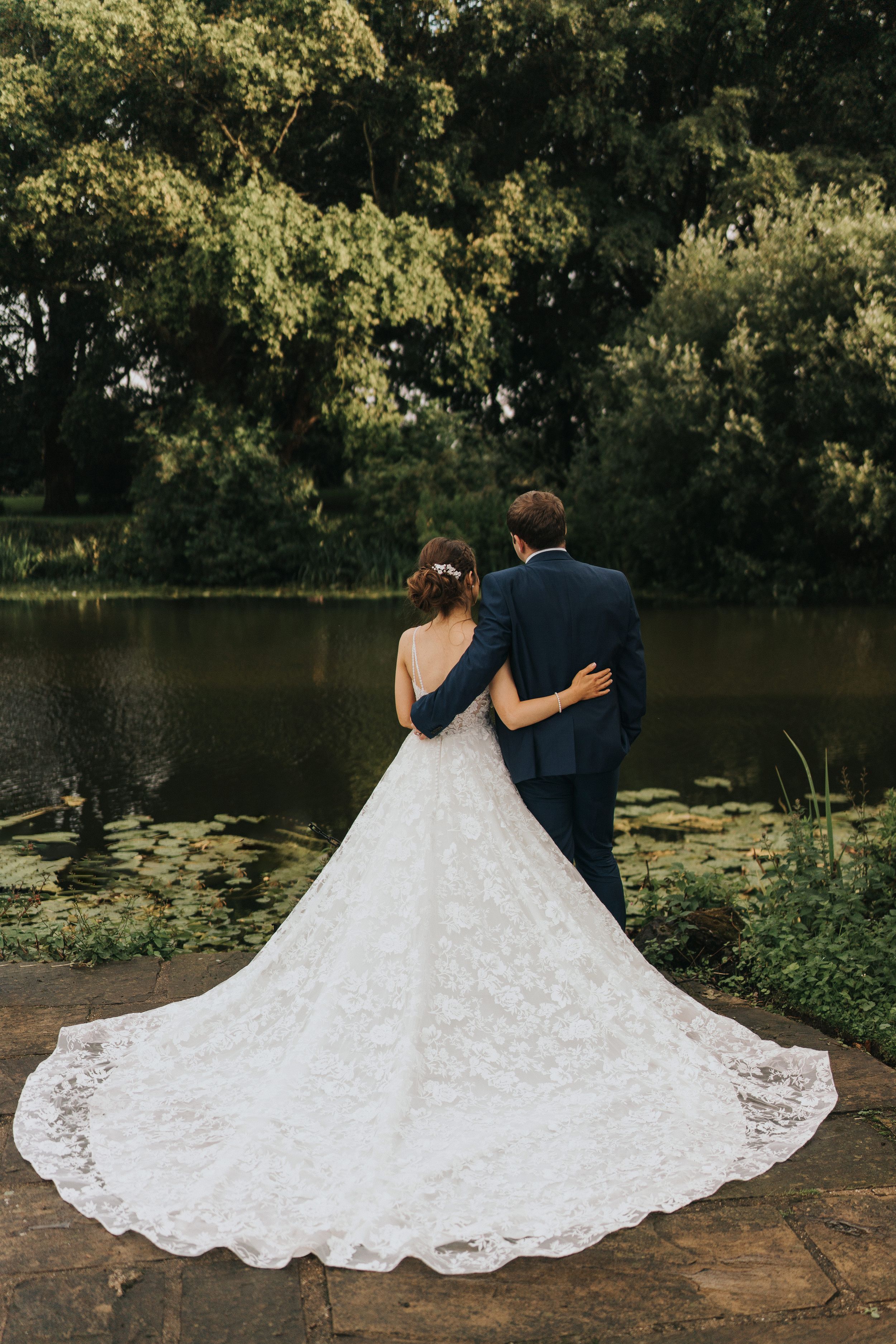 A newlywed couple stands at the edge of a tranquil pond, with the bride's elegant lace train flowing across stone ground at Cave Castle.