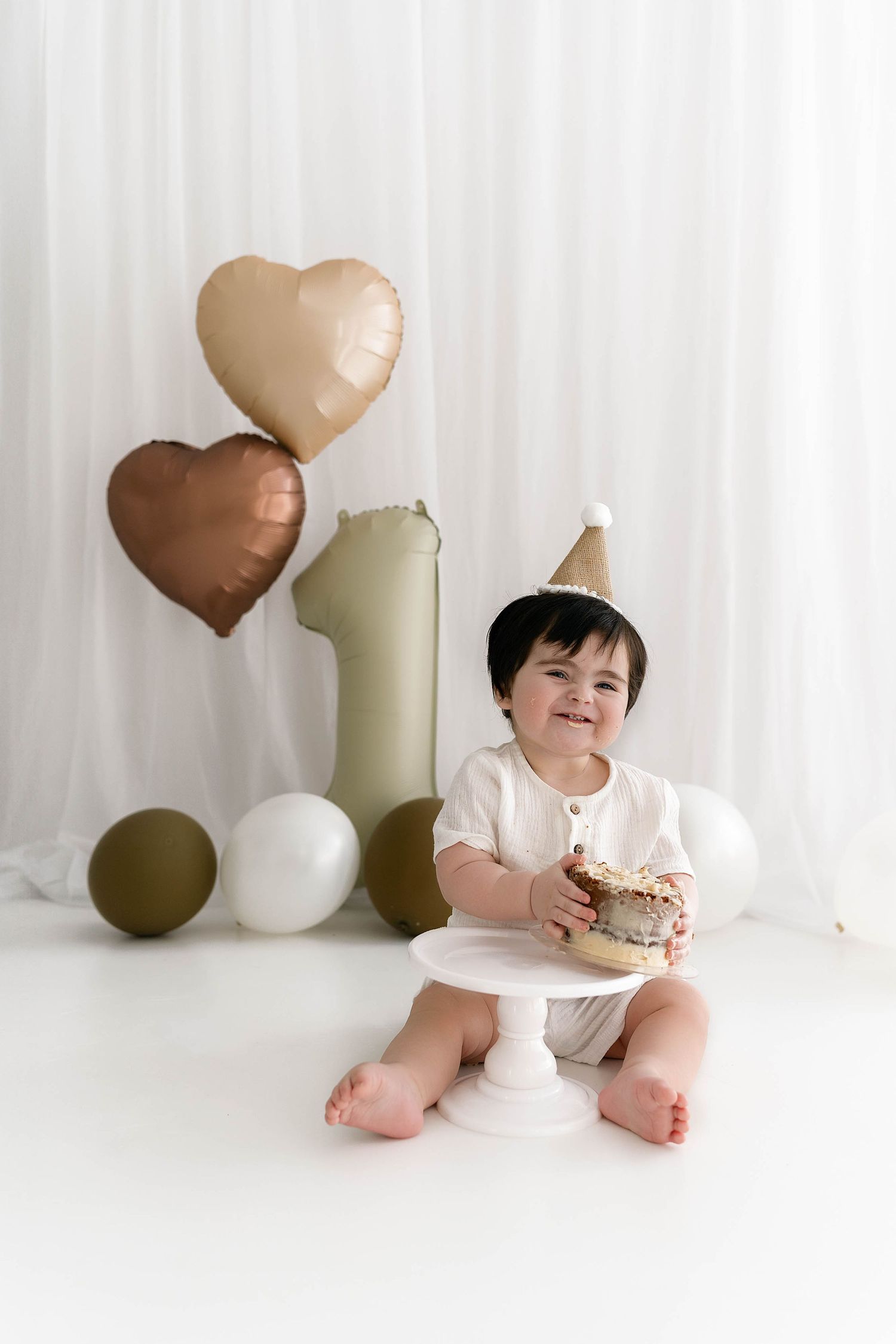 A baby in white outfit poses sweetly against white backdrop with heart-shaped balloons during a studio photo session.