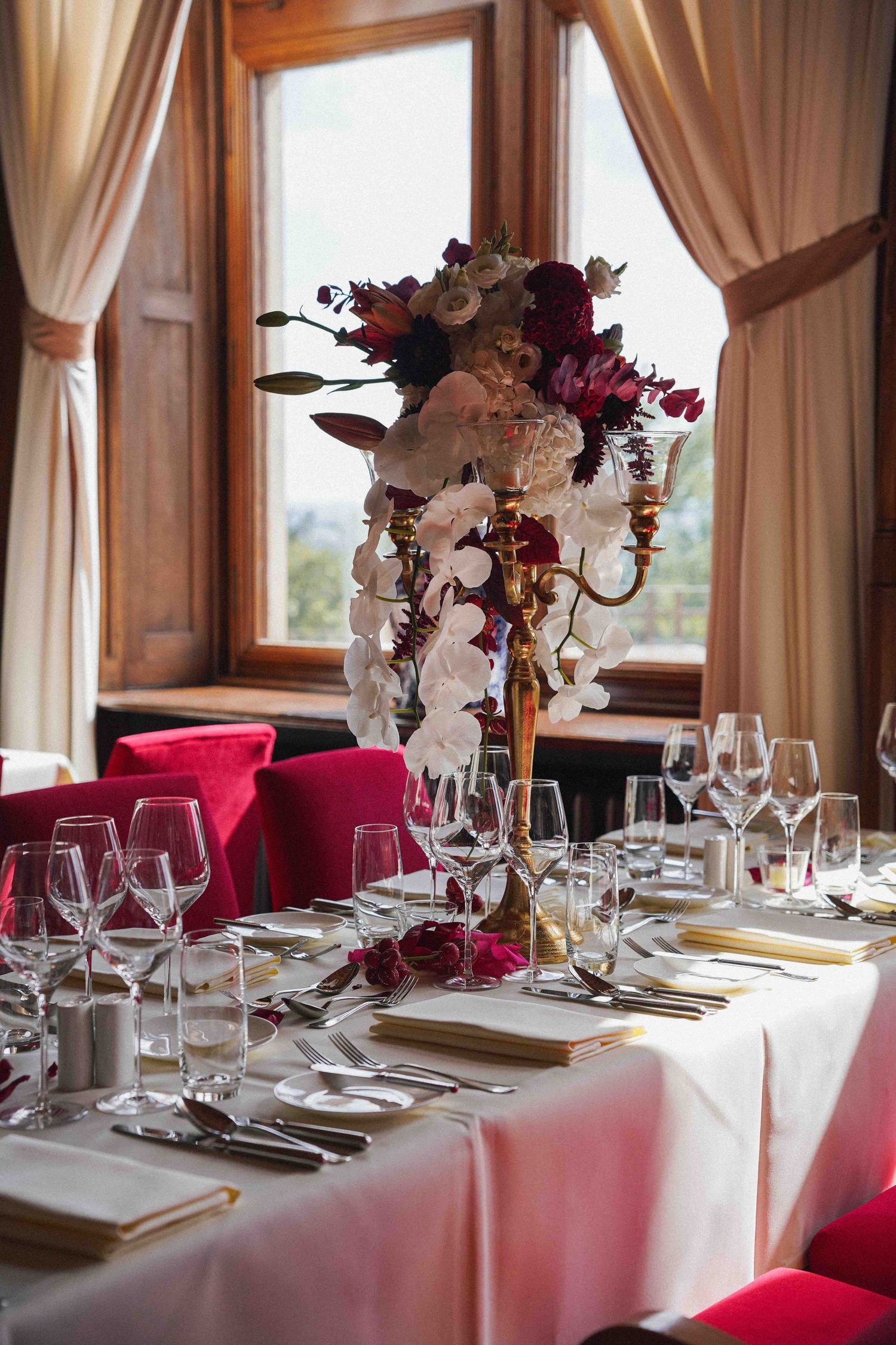 Elegant table setting with tall floral centerpiece, wine glasses and red velvet chairs in a sunlit dining room.