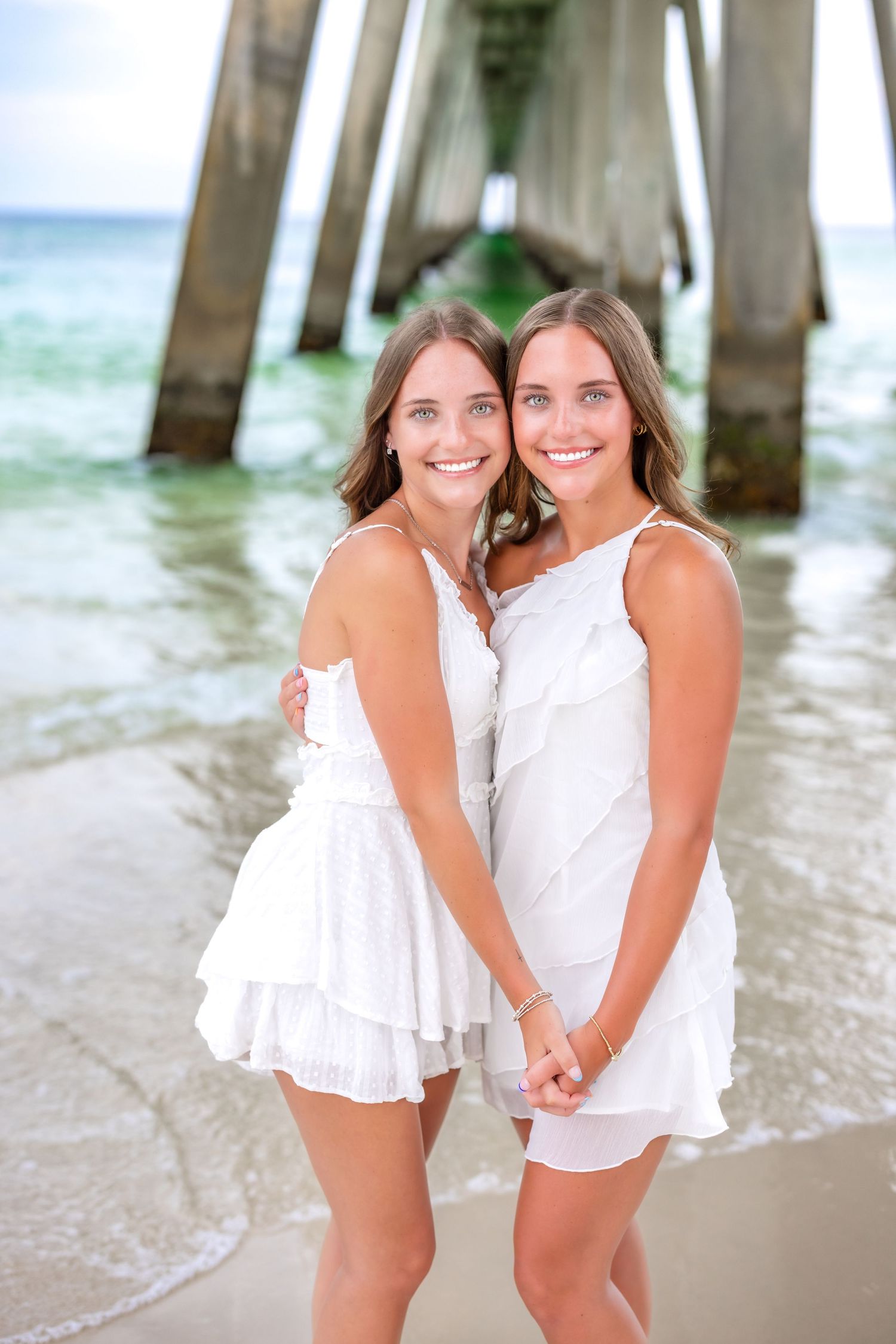 Two children in white dresses pose playfully together on a beach near a pier at sunset.