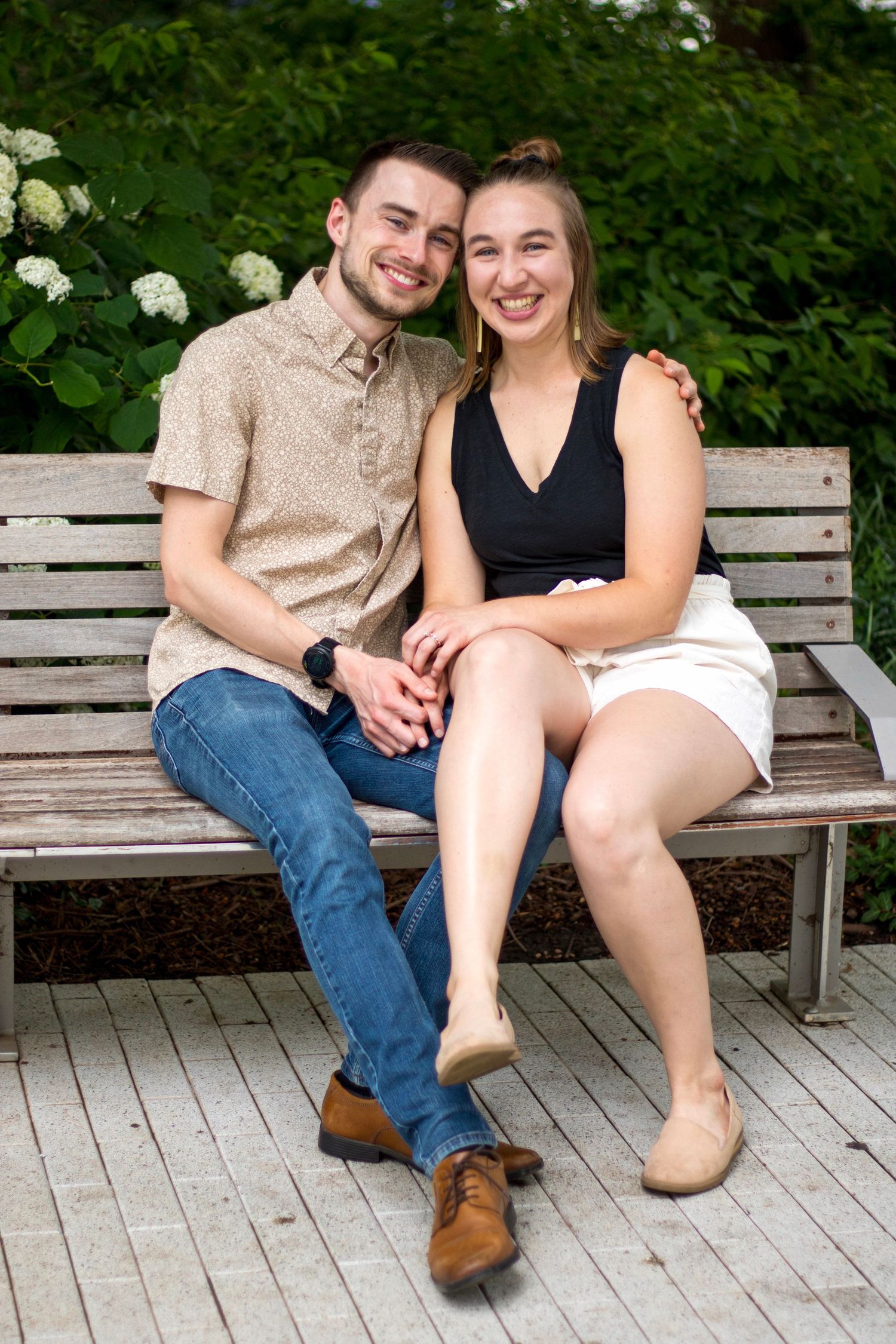A smiling couple sits closely together on a wooden bench surrounded by lush greenery, holding hands in a park.