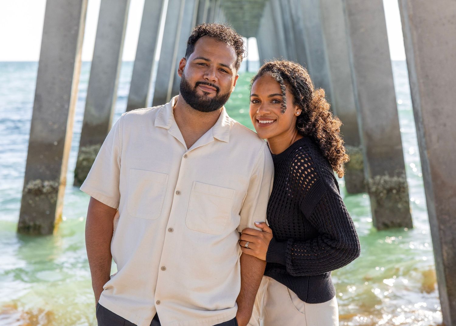 Happy couple posing together under pier by ocean in casual attire.