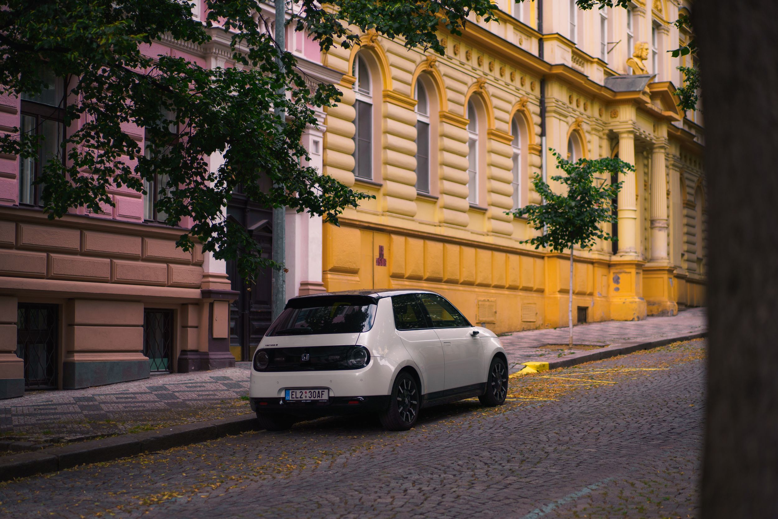 A white Honda E compact car parked on a slope on a leafy cobbled road with pastel pink and yellow buildings behind it