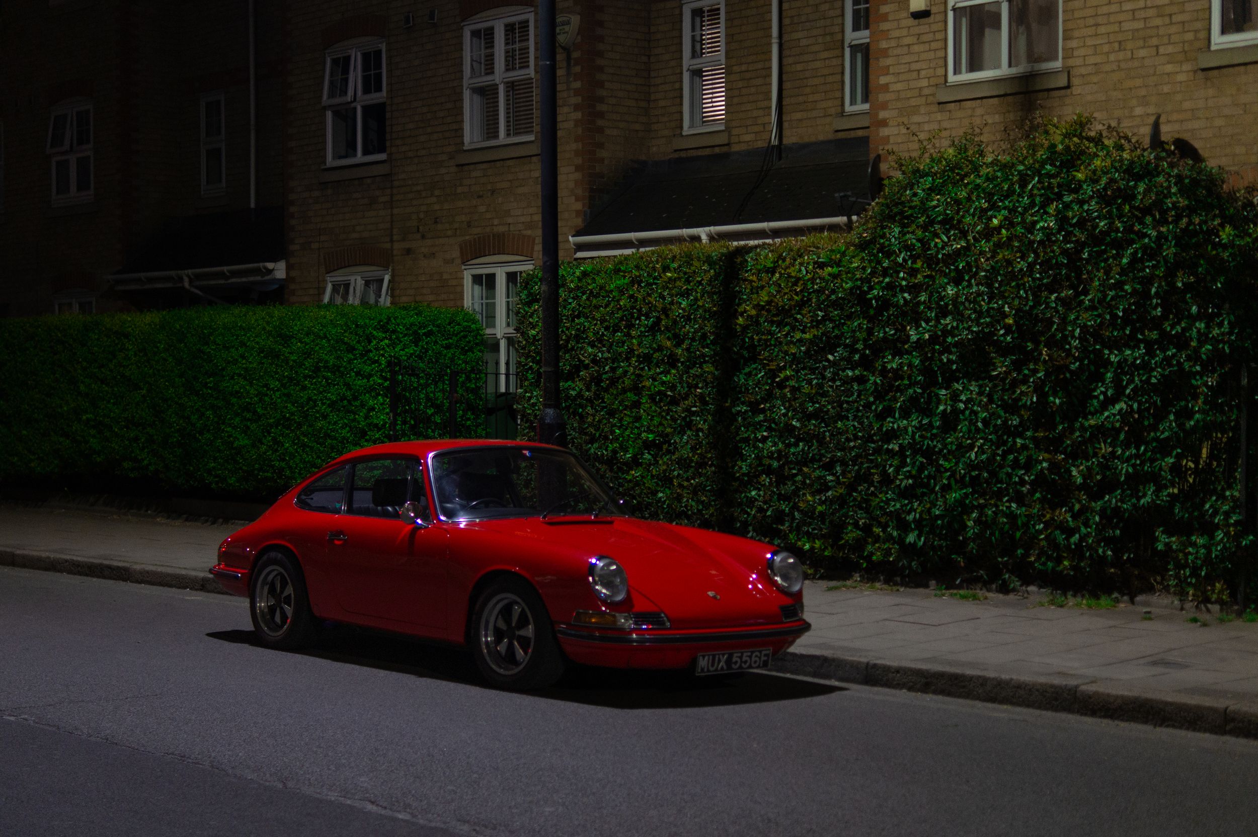 A red vintage Porsche car parked on the street at night with a hedge and houses behind it