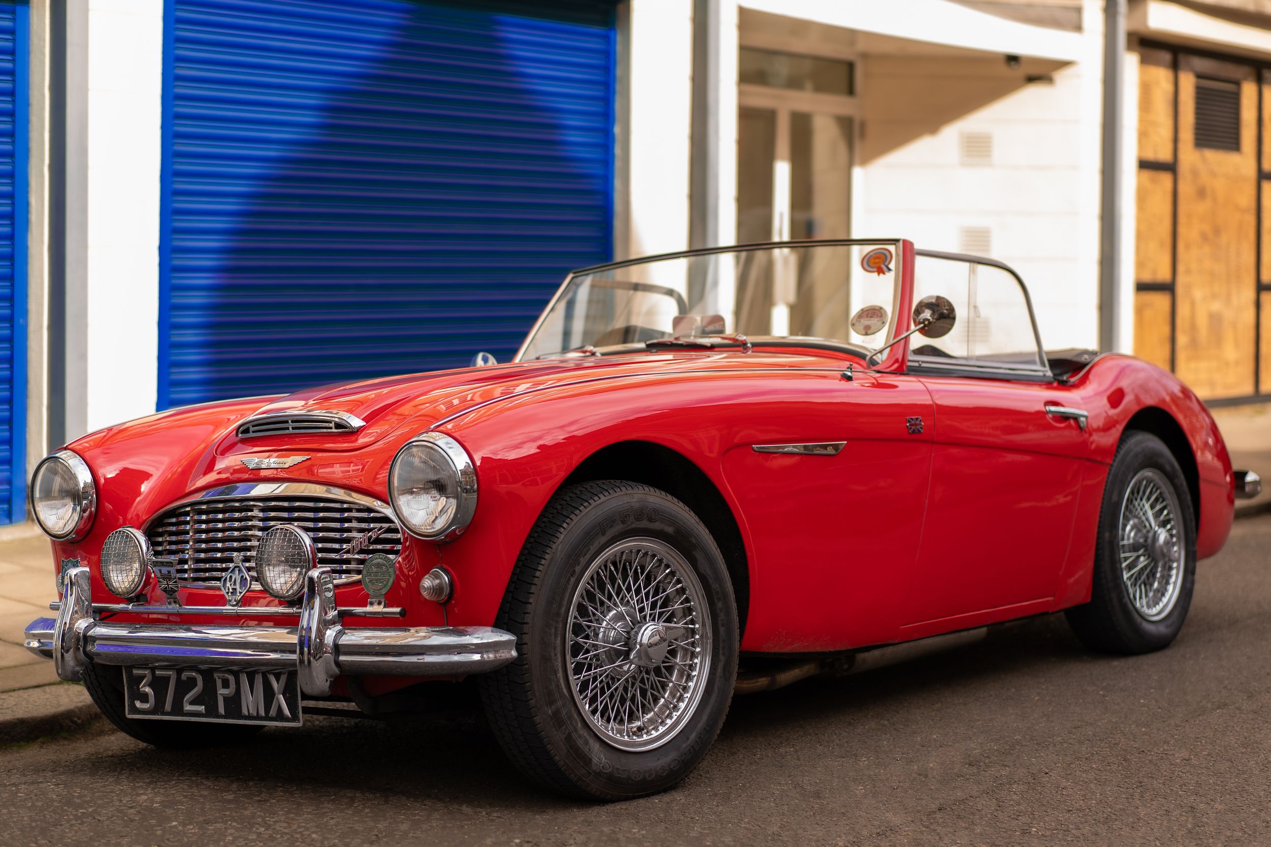 A red Austin Healy classic car with many association badges on the front bumper and a blue garage in the background