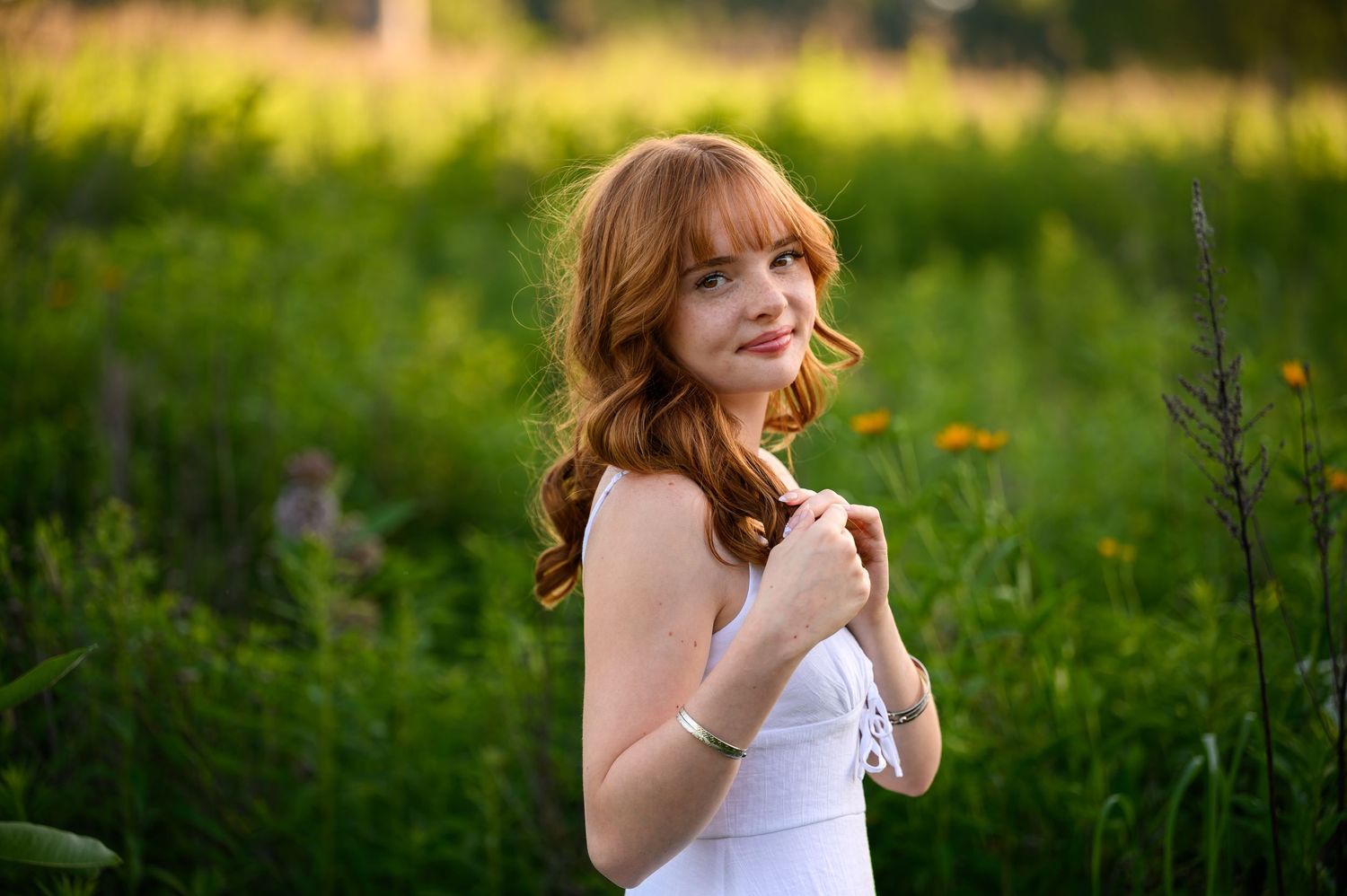 Young woman in white sundress poses outdoors in a lush green meadow with wildflowers at golden hour at The Highlands in Grand Rapids