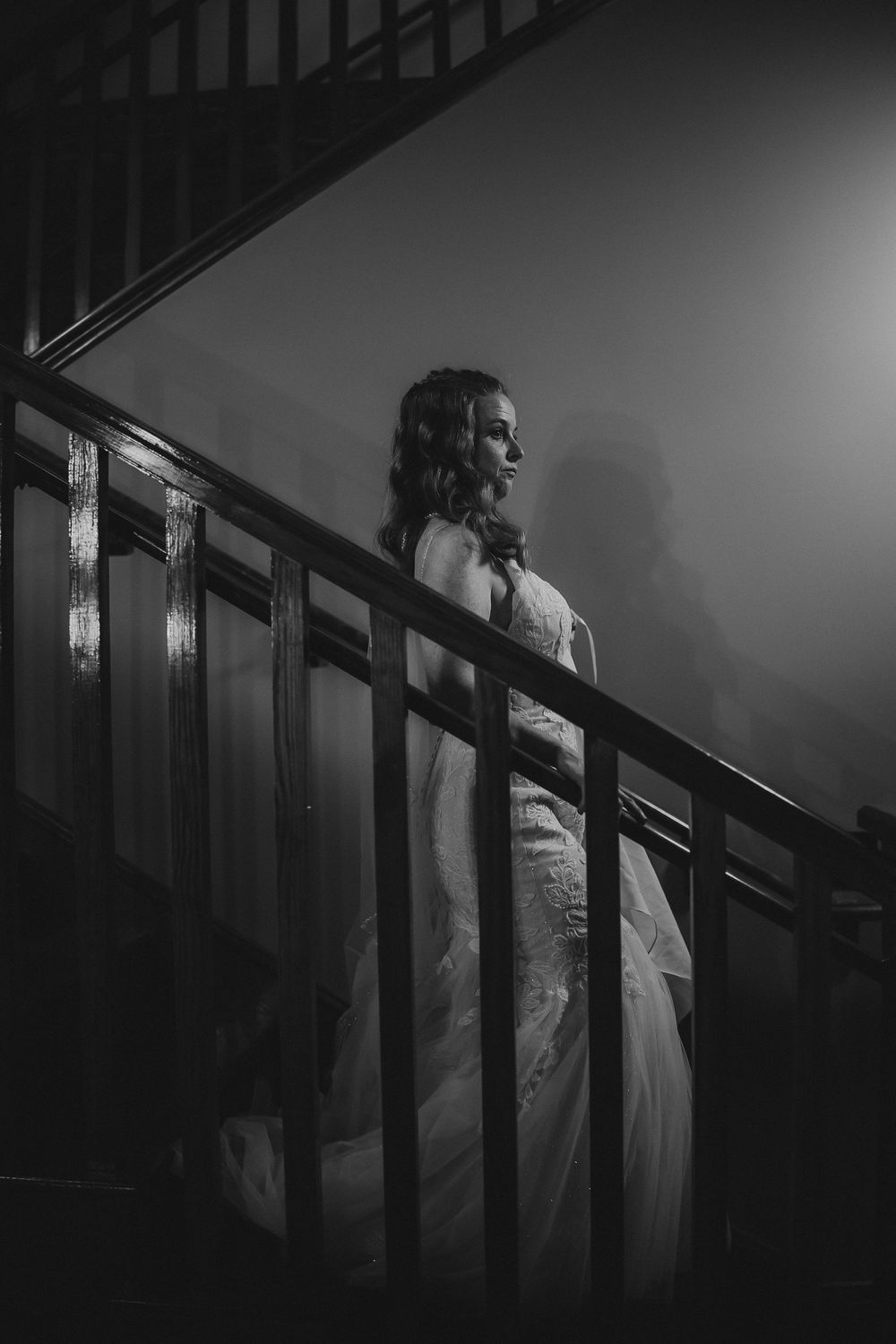 A figure in a white dress ascends a dark staircase casting dramatic shadows in black and white film noir style.