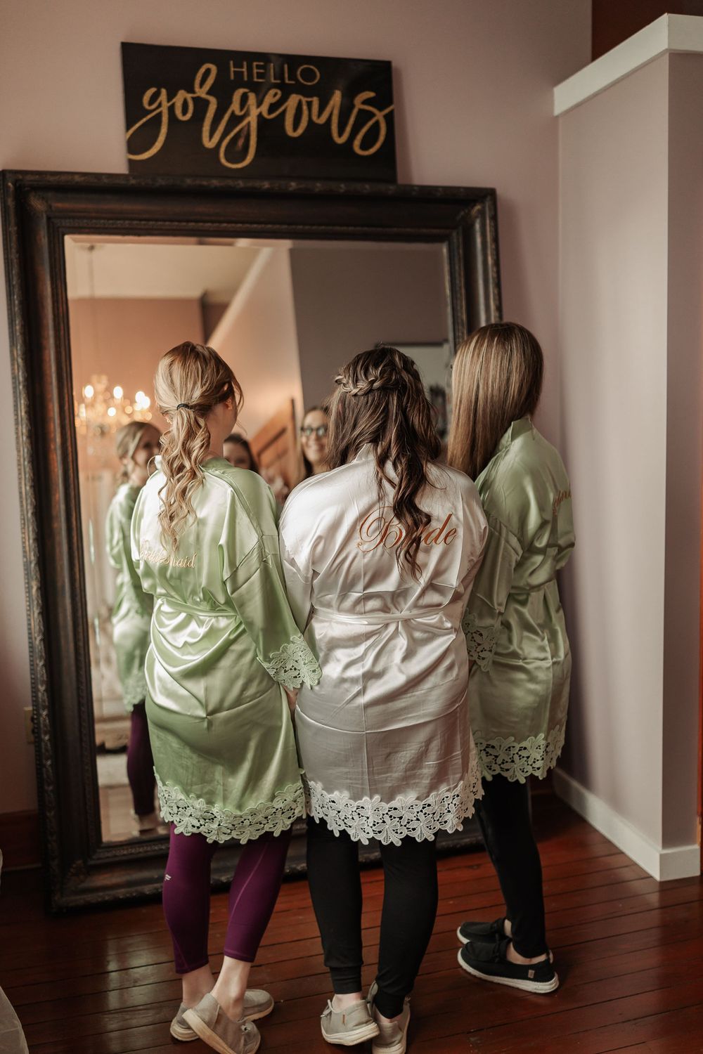 Three women in matching sage green floral lace robes looking at their reflection in a framed mirror below a rustic sign.