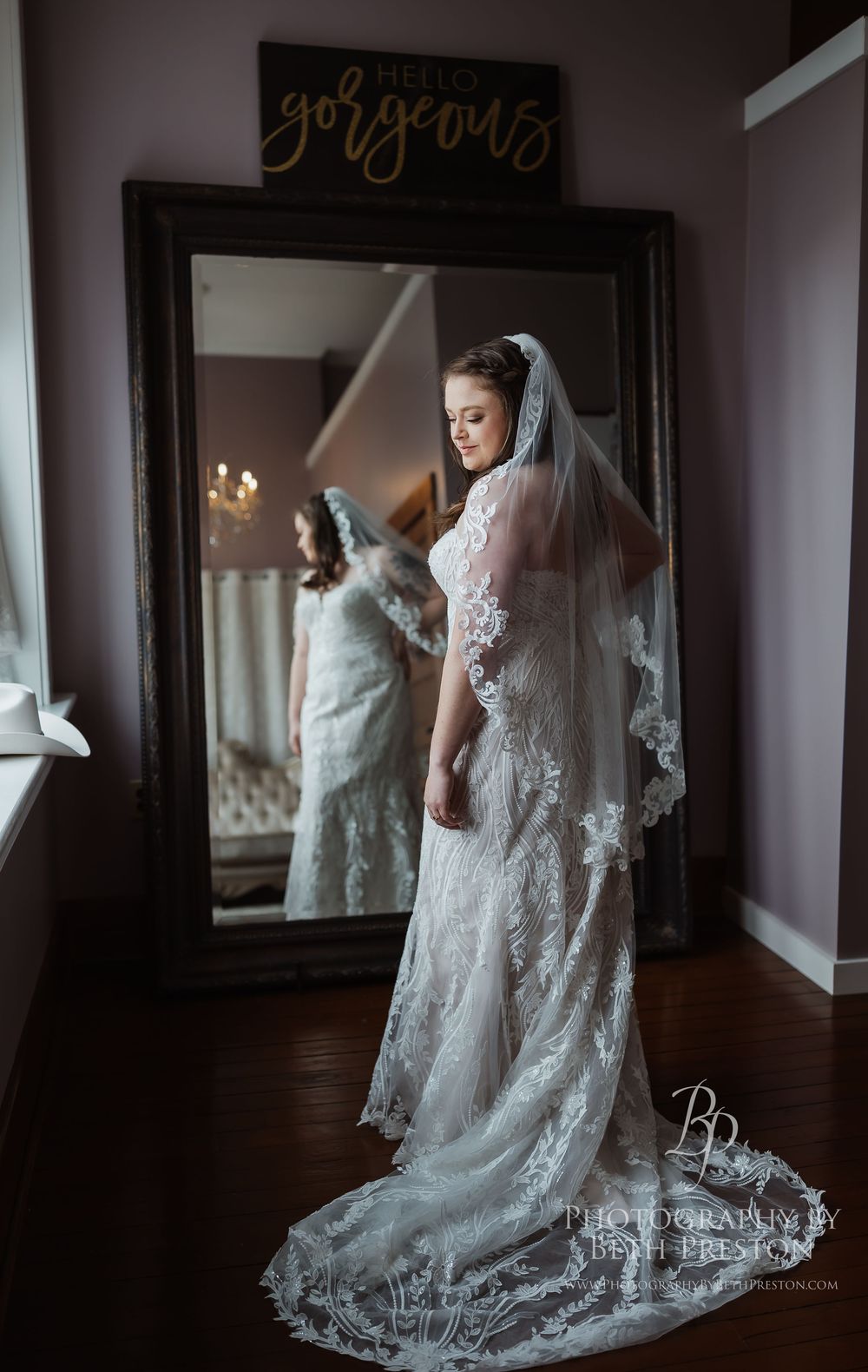 A bride in a white lace wedding gown with a long flowing veil stands elegantly against dramatic window lighting.
