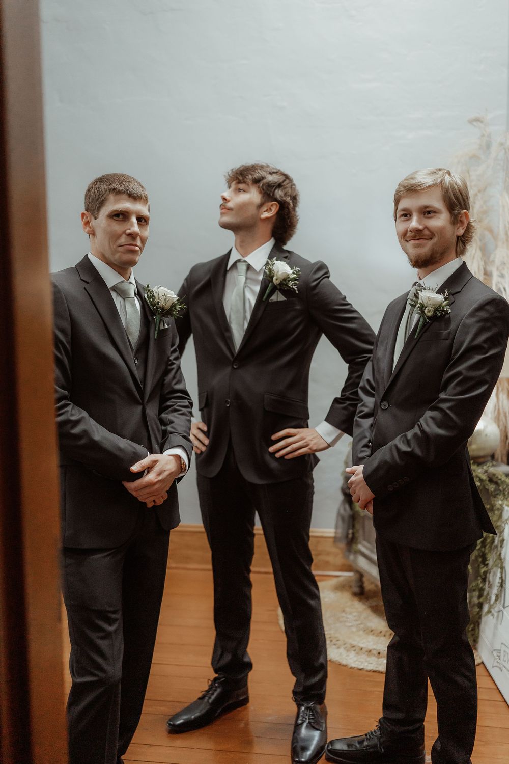 Groomsmen in black suits strike playful poses while getting ready for a wedding ceremony.