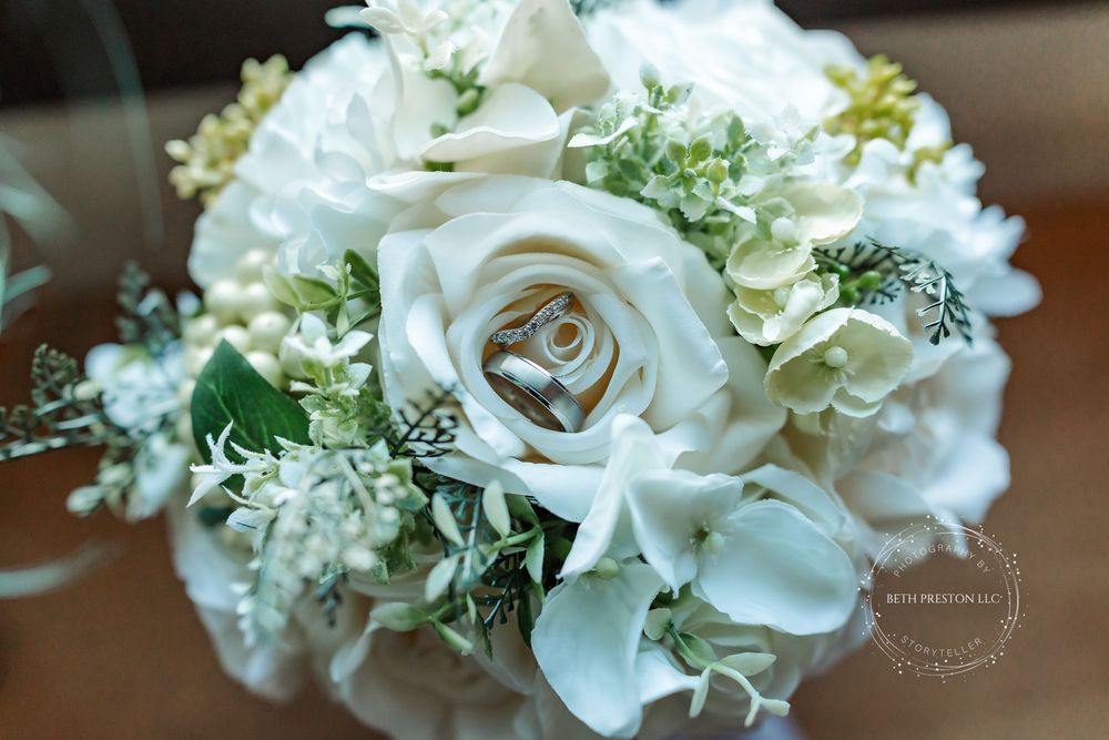 White rose bridal bouquet with wedding rings resting on top, featuring hydrangeas and greenery.
